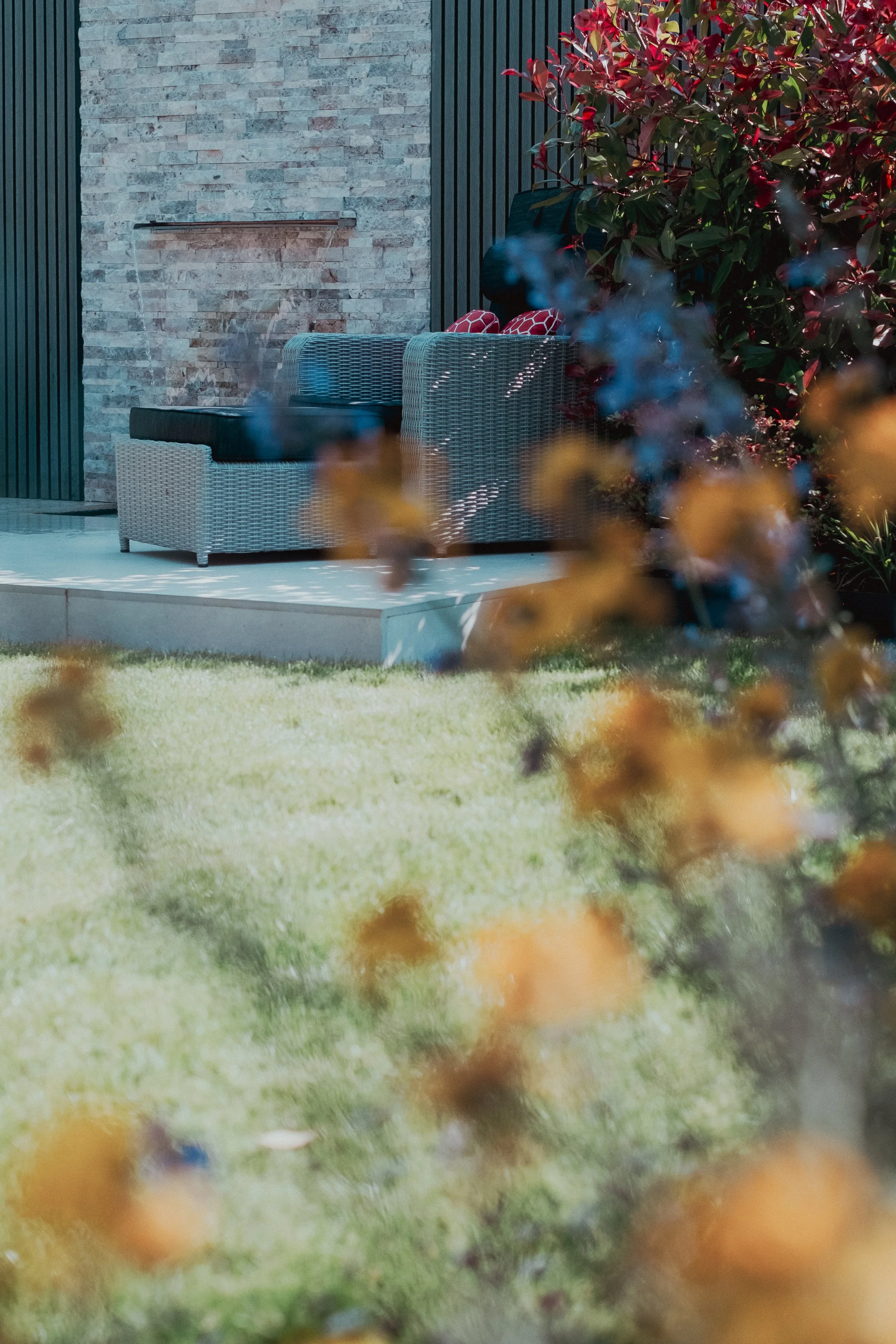 Outdoor patio with wicker sofa, brick and wood wall, surrounded by plants and grass.