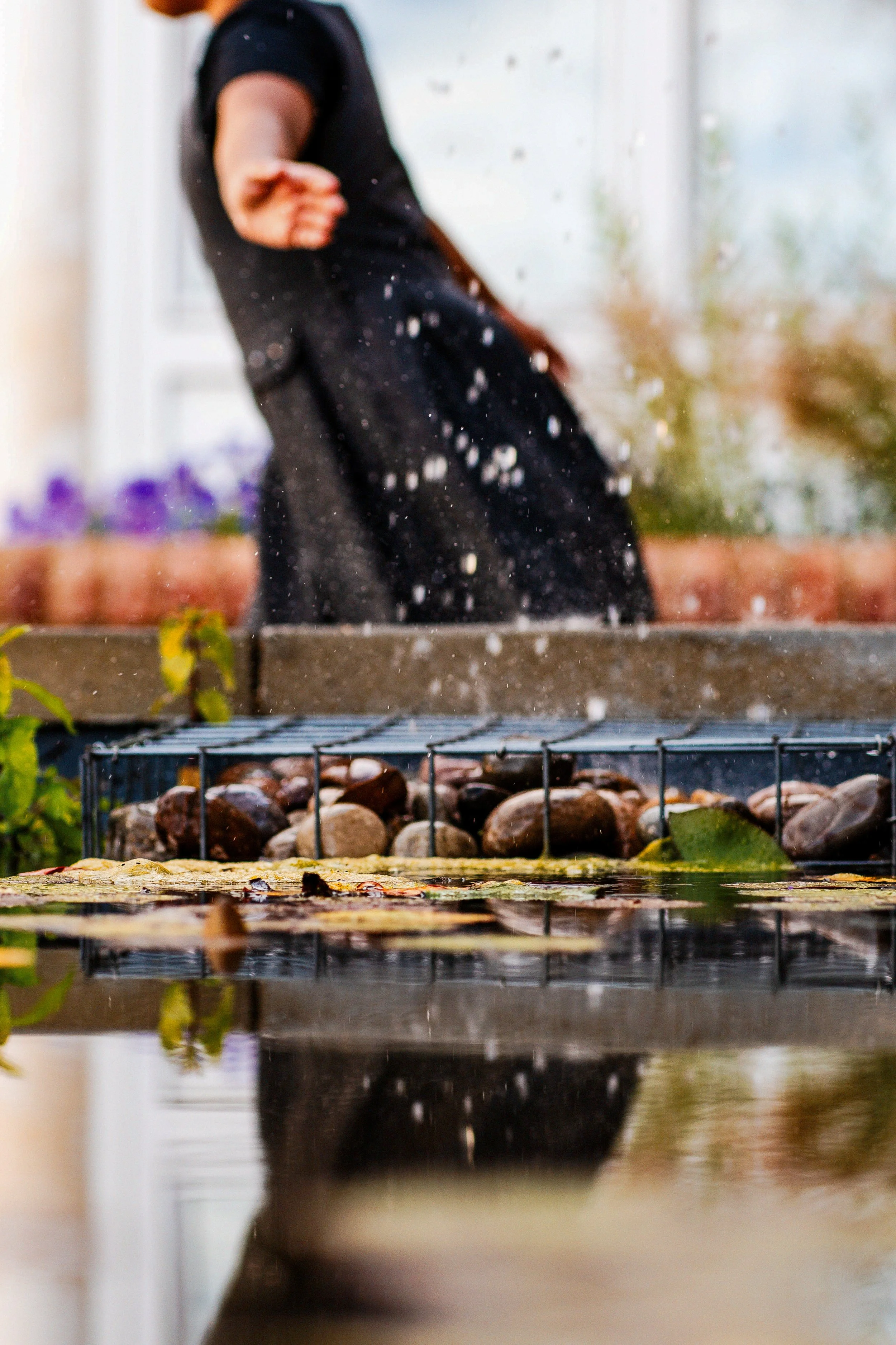 A person in a black dress splashing water outdoors on a day with cloudy weather, with a pond, stones, and plants visible in the foreground.
