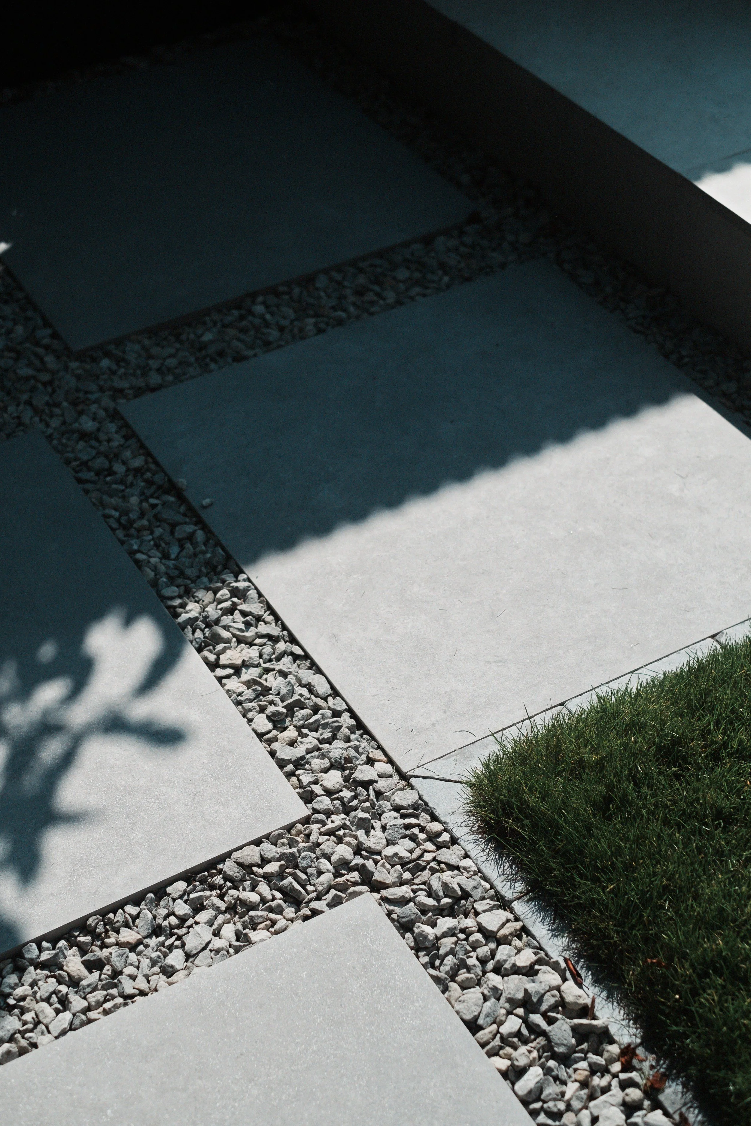 Close-up of modern outdoor pathway with large square concrete tiles and gravel-filled gaps, partially shaded, with a patch of grass in the corner.