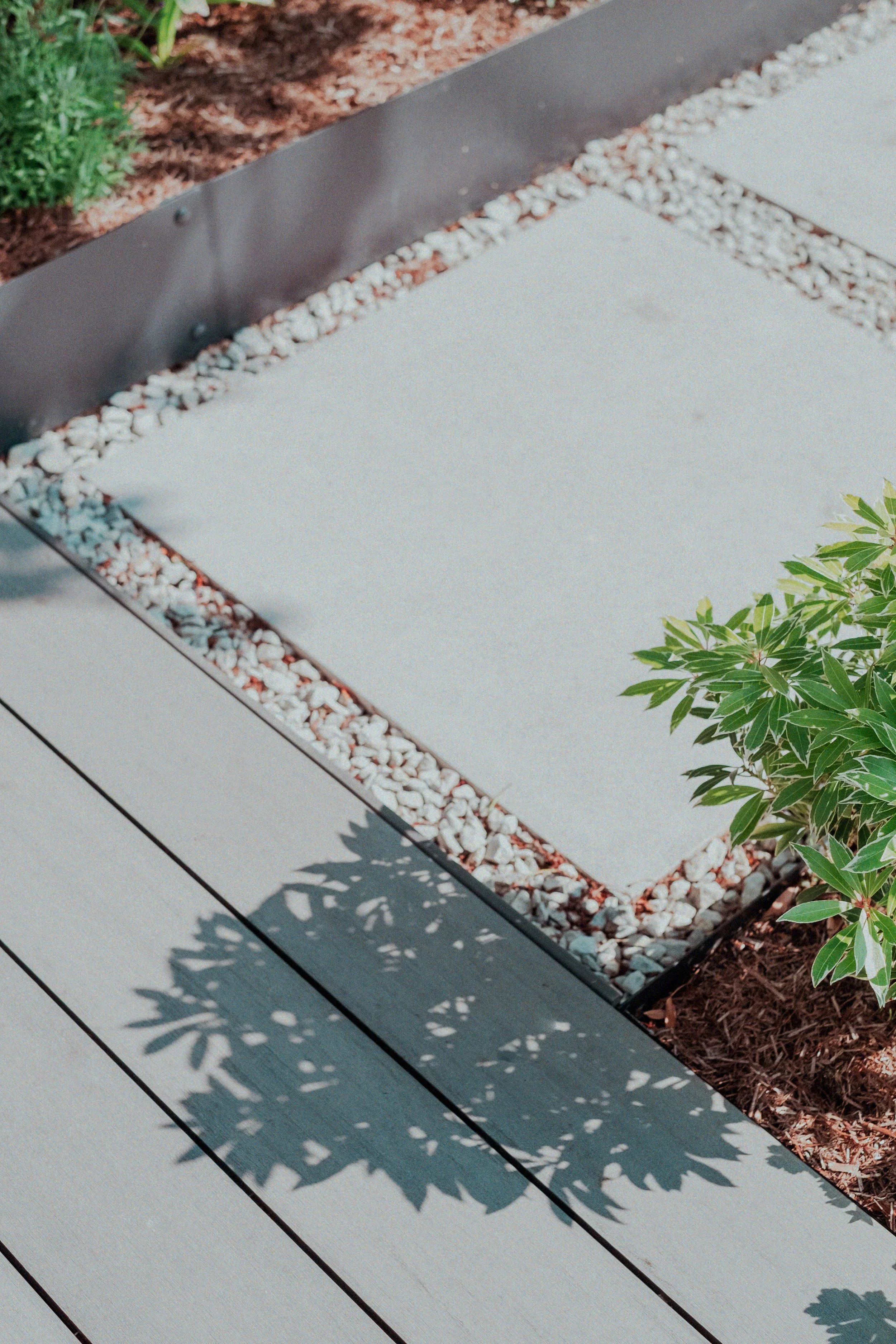 Close-up of concrete pathway with gravel edges and a shadow of a leafy plant on the ground, adjacent to a garden bed with green foliage.
