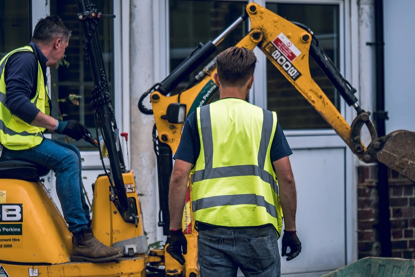 Two workers in safety vests operating a small backhoe loader outside a building.