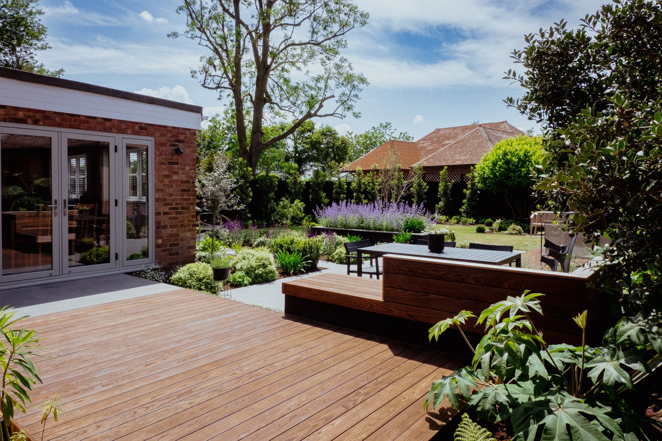 A backyard garden with a wooden deck, outdoor furniture, lush green plants, flowers, trees, and a house with a brick wall and sliding glass doors.