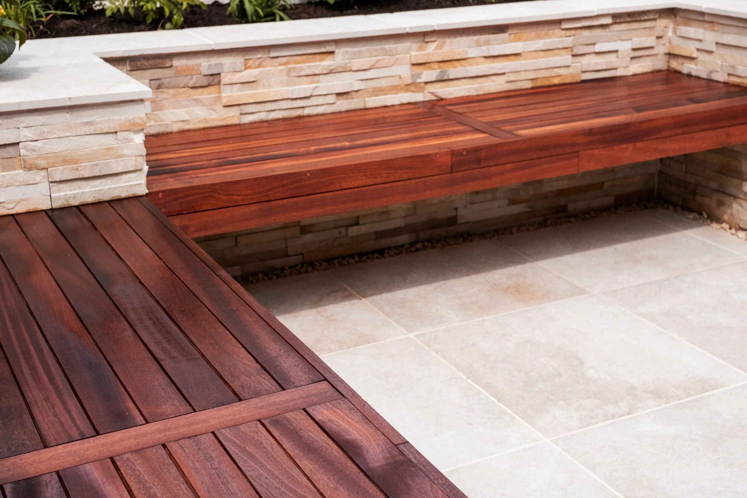 A section of outdoor space featuring a built-in wooden bench made of polished wood planks, with a surrounding wall of stacked stone bricks and a tiled floor.
