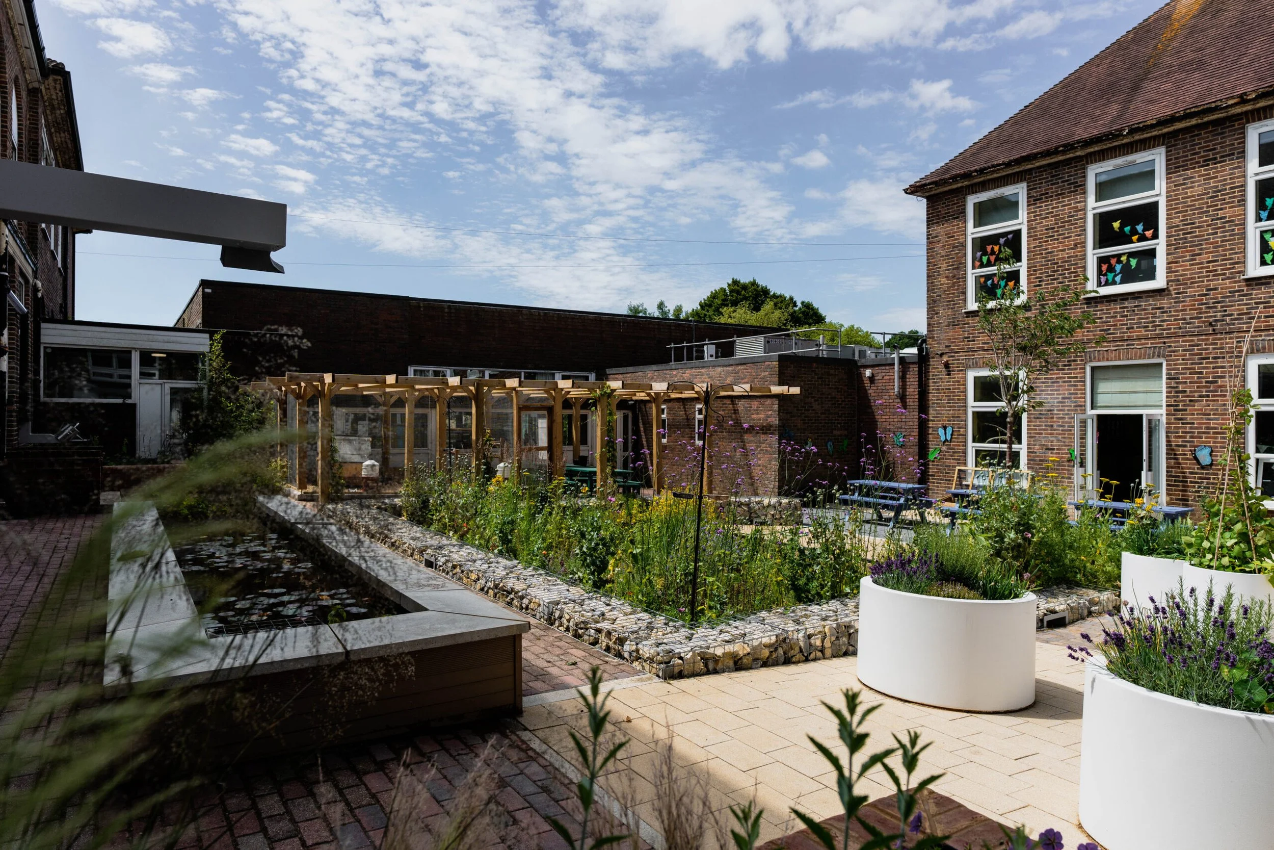 A garden courtyard with a raised flower bed, potted plants, and a brick building in the background, under a partly cloudy sky.