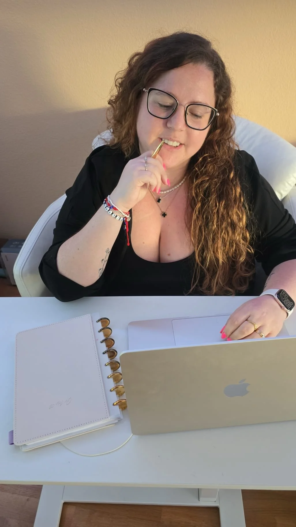 Mulher com cabelos cacheados, usando óculos, brincando com um bolígrafo, sentada em frente a um notebook prata de maçã, com uma agenda branca ao lado, em um ambiente interno