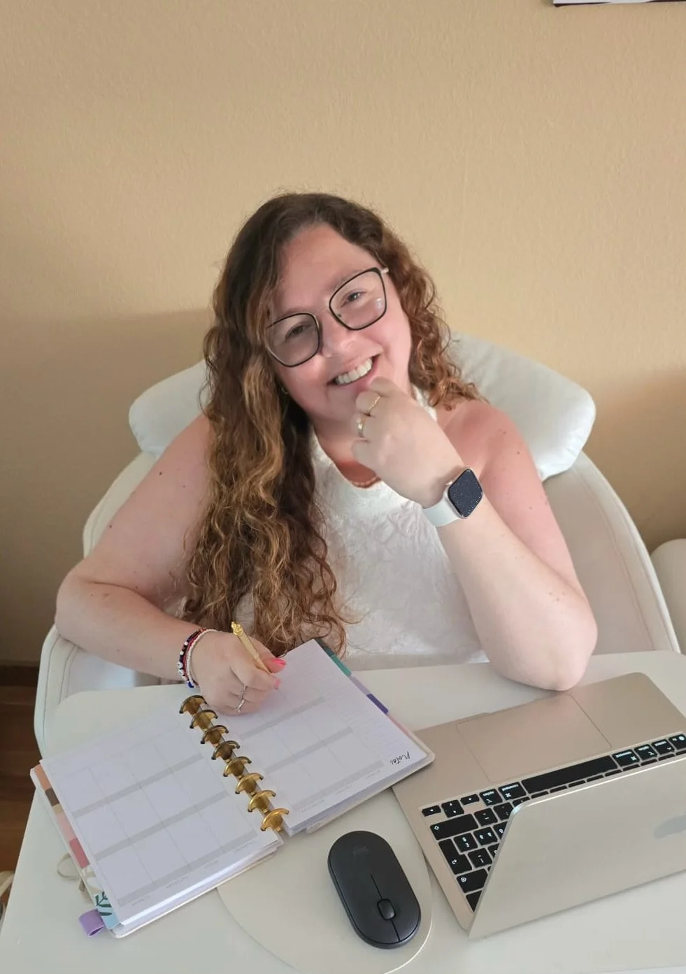 Mulher sorridente com cabelo cacheado, óculos e vestindo branco, sentada na mesa com um planner, laptop, mouse e usando uma smartband, em um ambiente interno