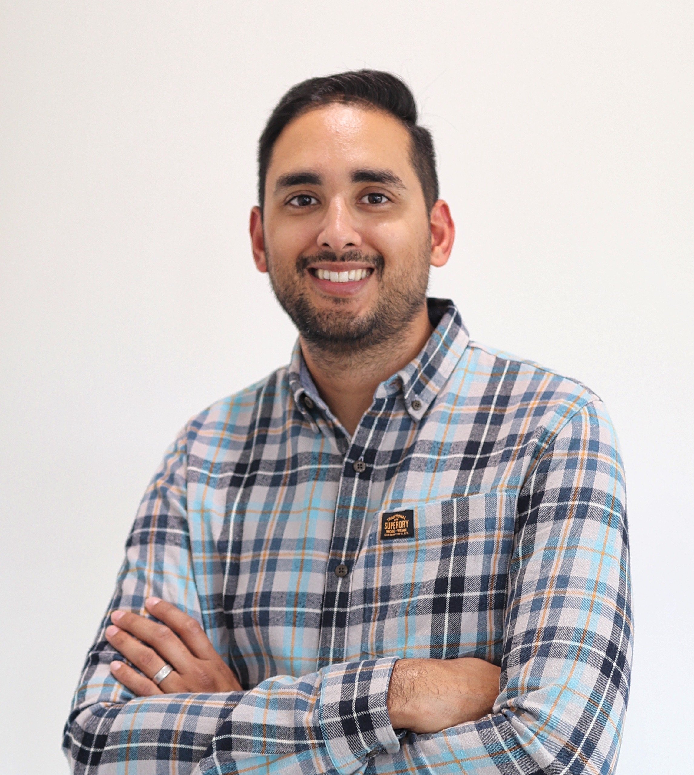 A man with dark hair and a beard, smiling, wearing a plaid button-up shirt, standing with arms crossed against a plain white background.
