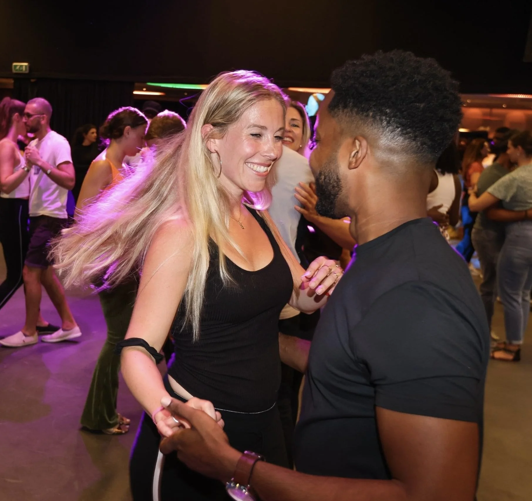 A smiling woman with long blonde hair dancing closely with a man in a dark t-shirt at a lively dance party with other people dancing in the background.