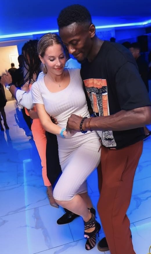 A couple dancing at a party with blue lighting. The woman is wearing a white dress and heels, and the man is wearing a black shirt and brown pants. They are close together, smiling and enjoying the dance.