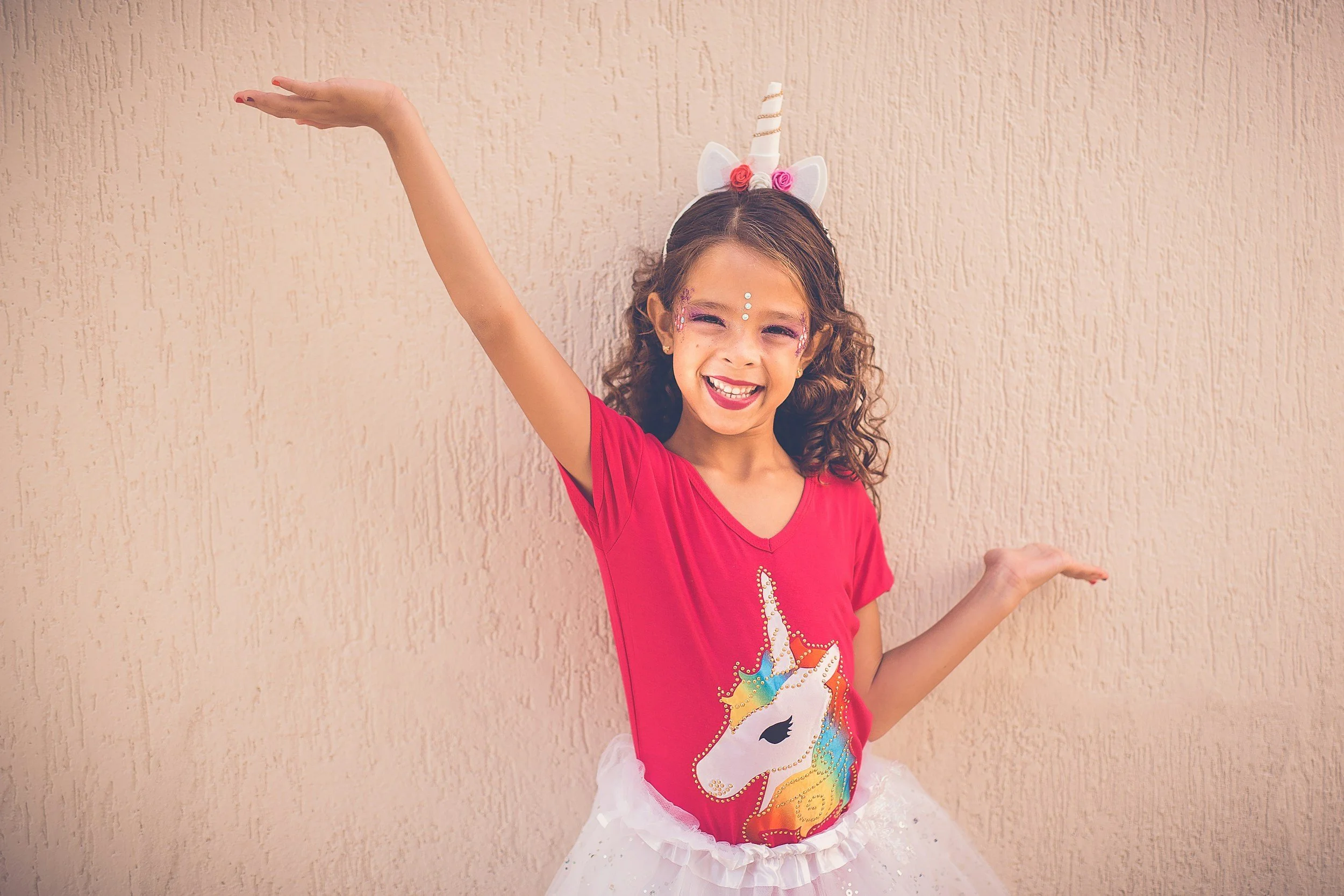 Young girl smiling in a unicorn-themed costume, standing against a beige textured wall.