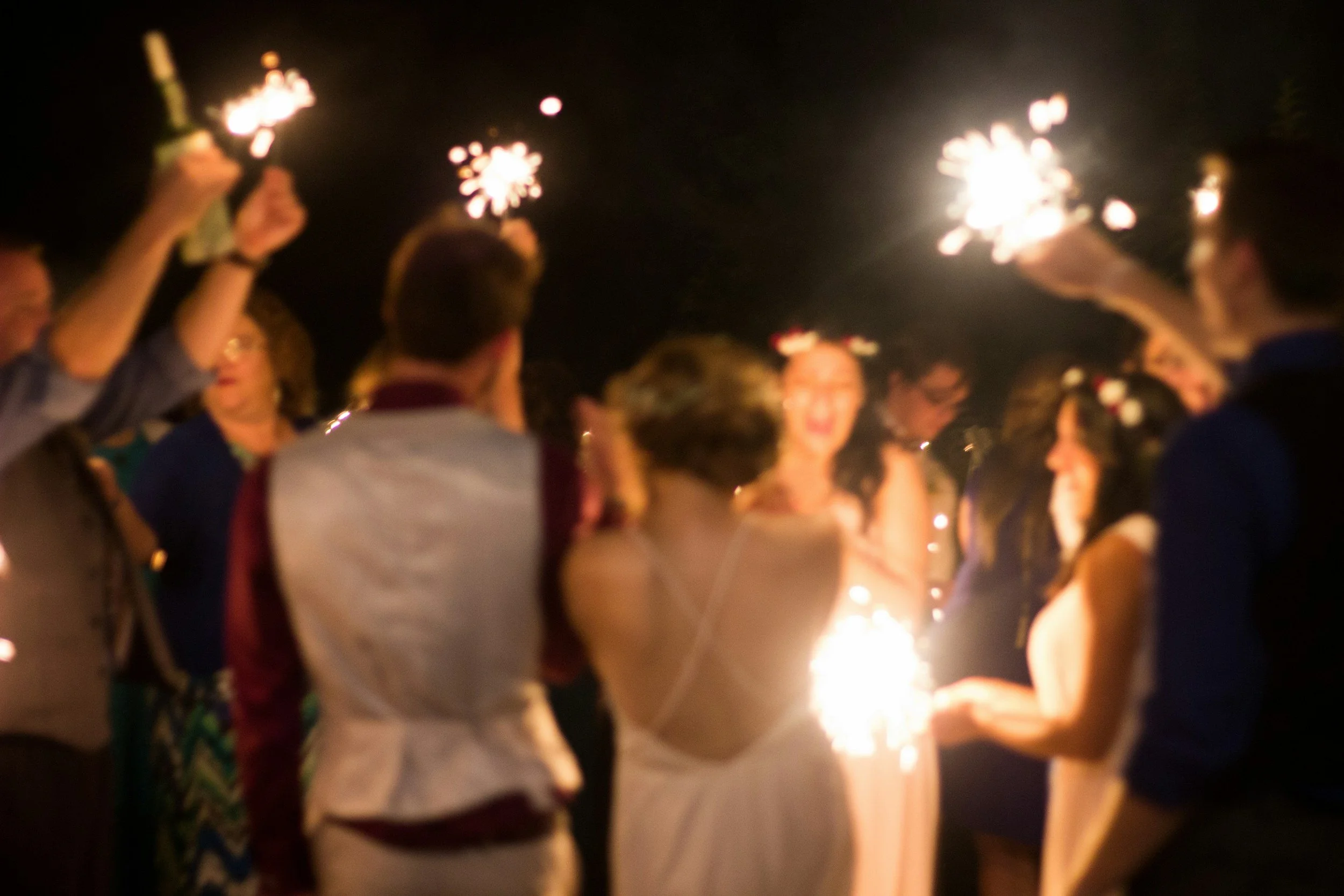 People celebrating at night with sparklers, some wearing flower crowns, smiling and talking.