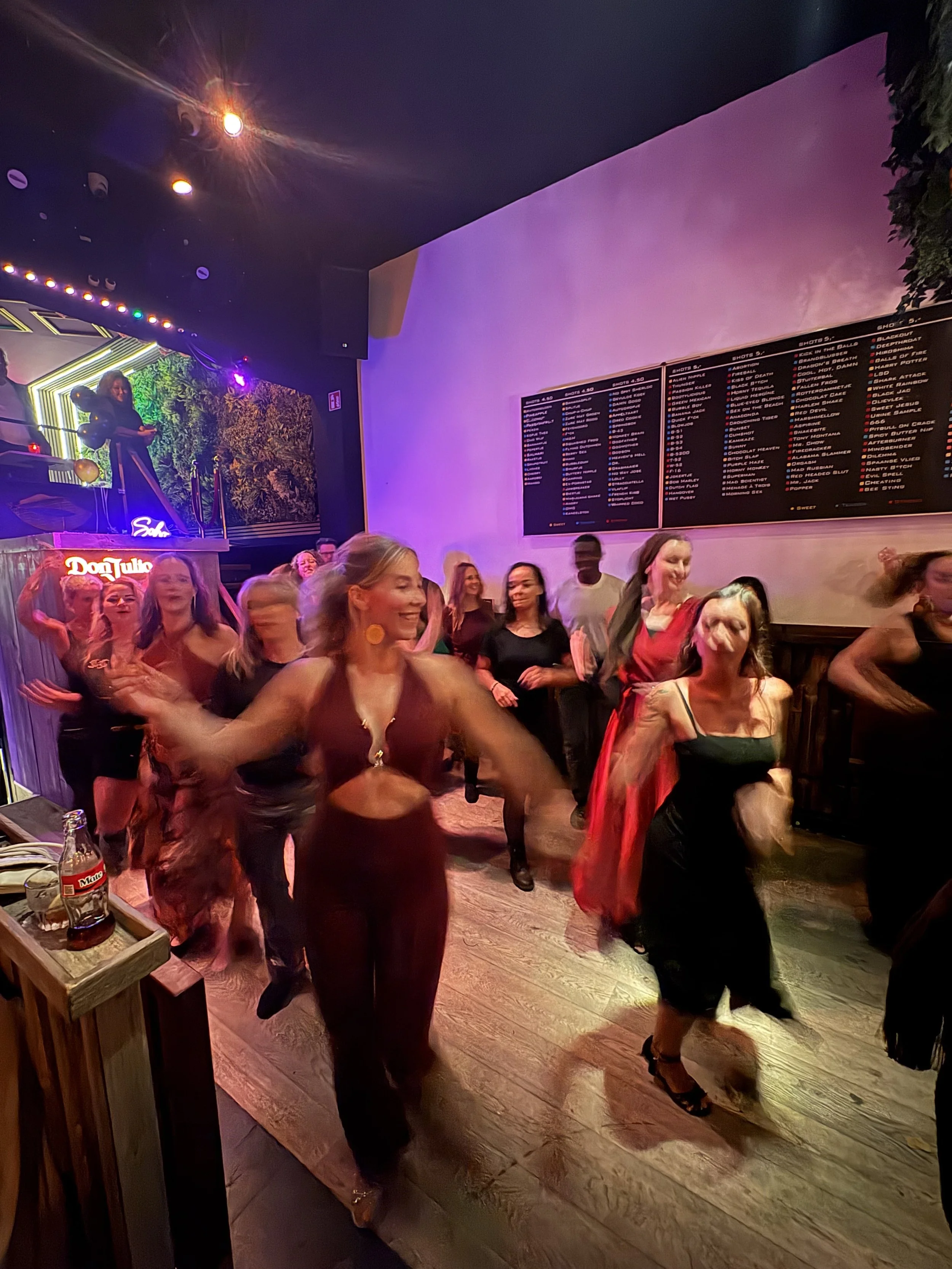 People dancing at a lively nightclub with colorful lighting, a large screen on the wall, and a woman holding balloons on the DJ booth.