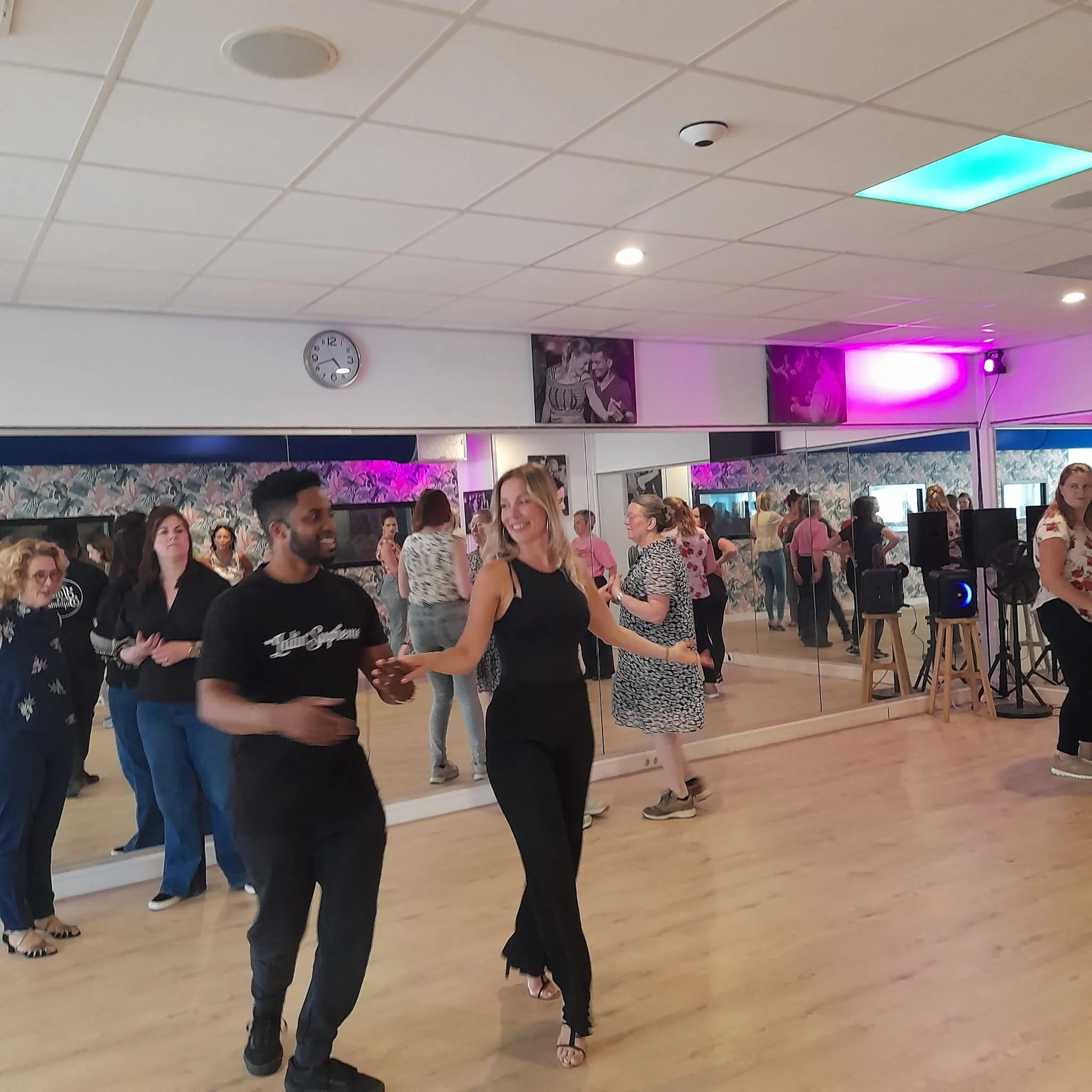 People dancing in a mirror-lined dance studio with colorful lighting.