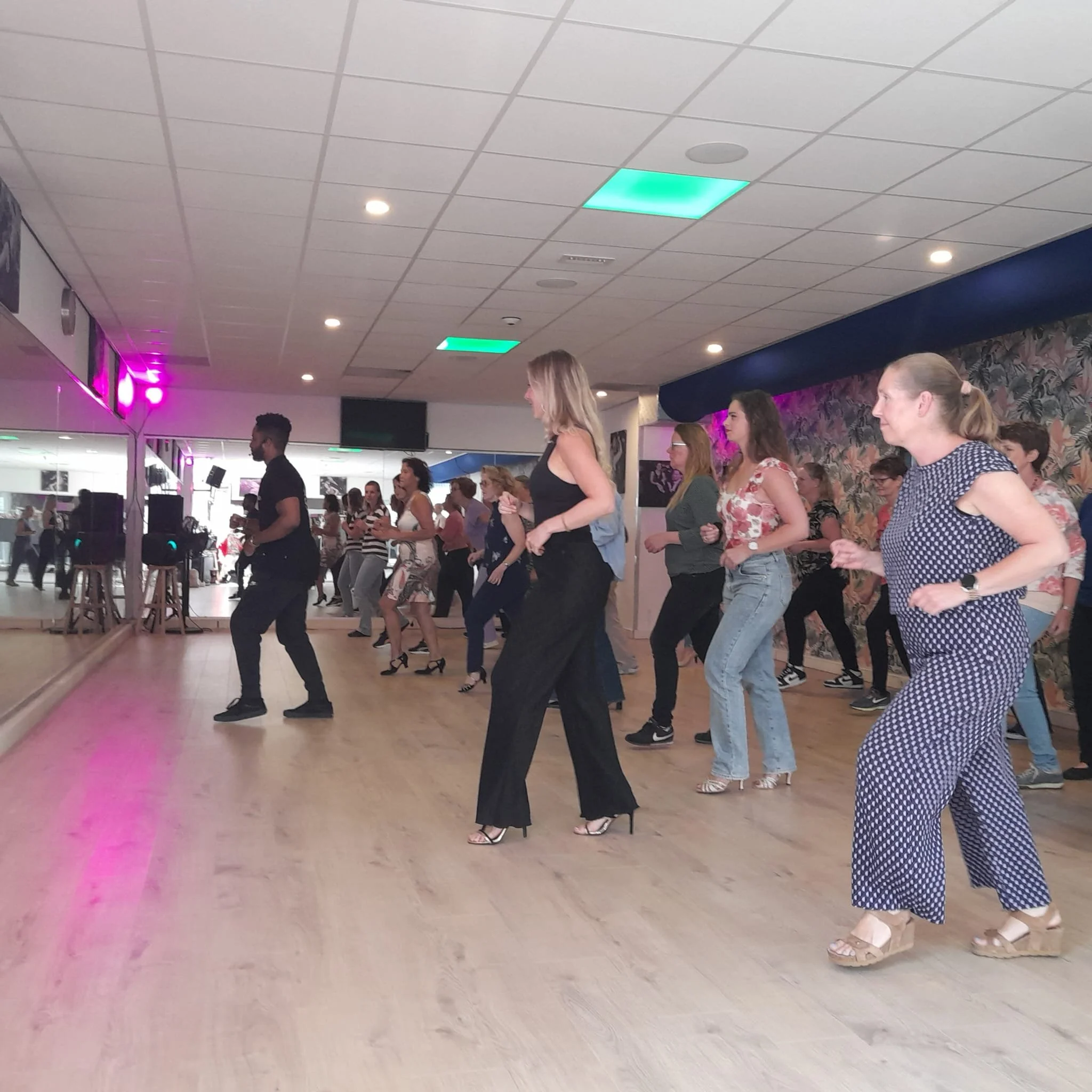 Group of people dancing in an indoor dance studio with a mirror wall and colorful ceiling lights.