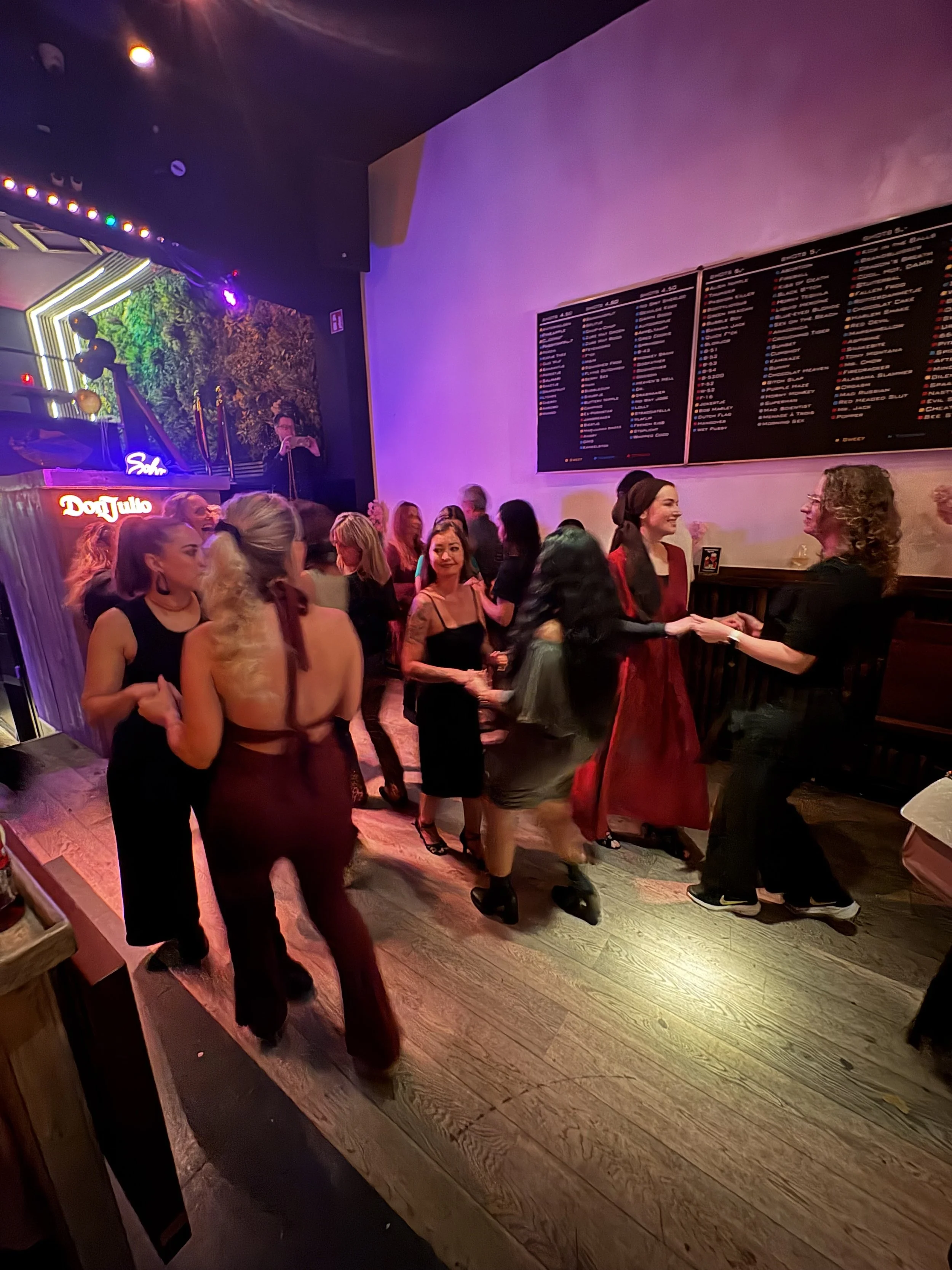 People dancing and socializing in a nightclub with colorful lighting, a DJ booth, and a large menu board on the wall.