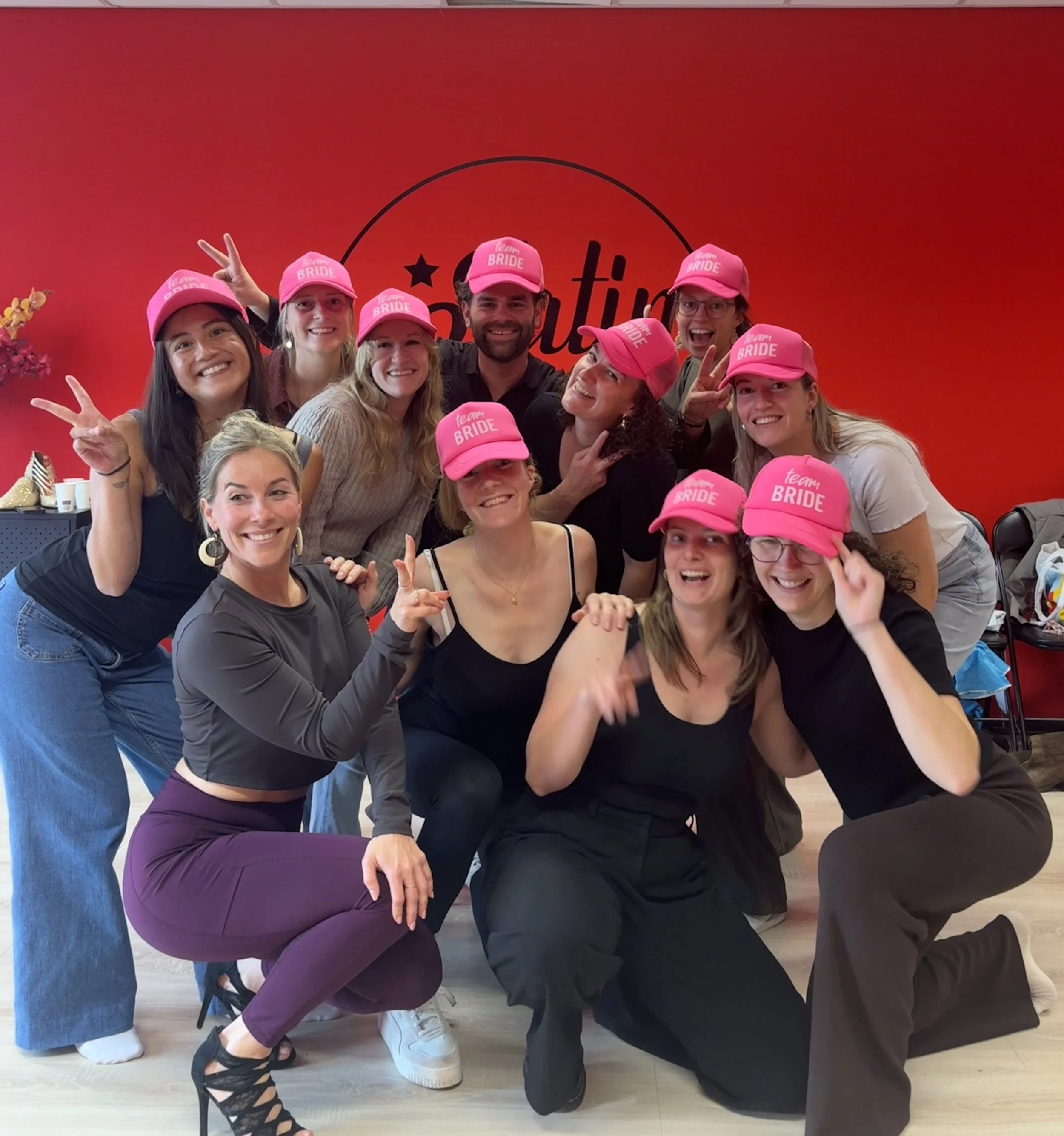Group of women and one man celebrating at a bridal shower, all wearing pink caps with "Team Bride" written on them, standing in front of a red wall with a partial logo that includes a star.