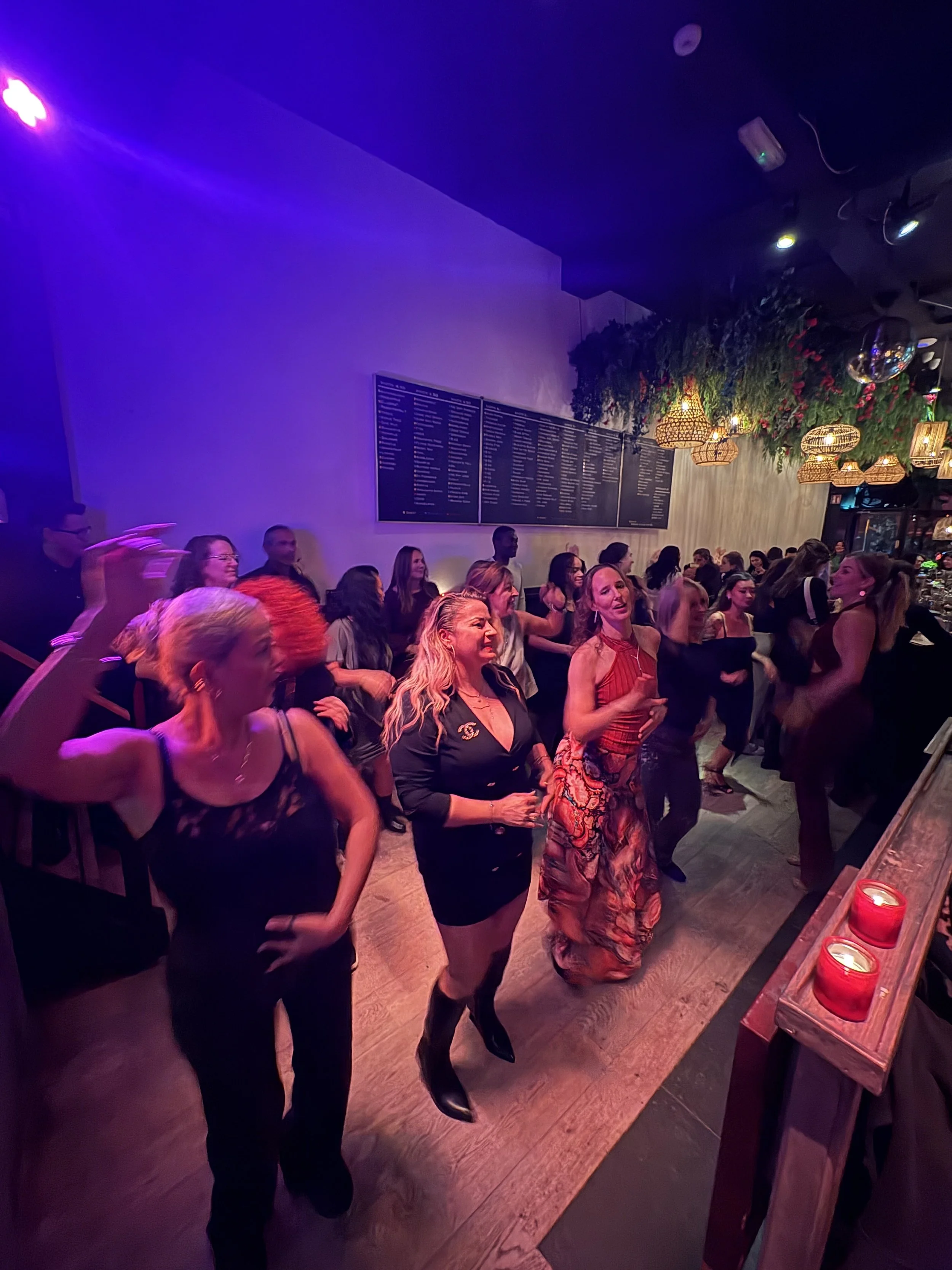 People dancing and enjoying music in a dimly lit bar or nightclub with purple and pink lighting, hanging lamps, and candles on a counter