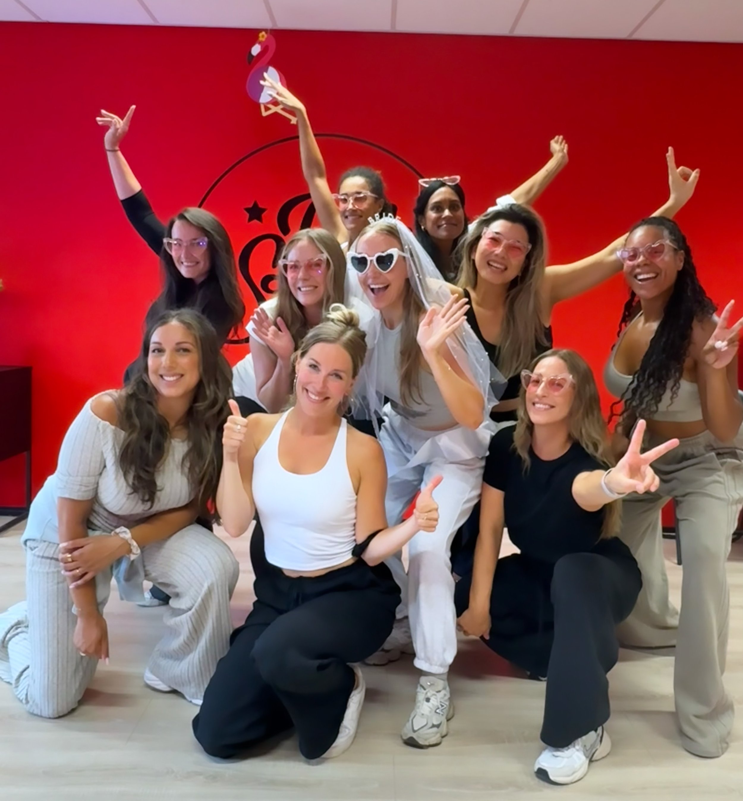 Group of women celebrating in front of a red wall, some wearing fun glasses, a bride with a veil, and making peace signs and thumbs up.
