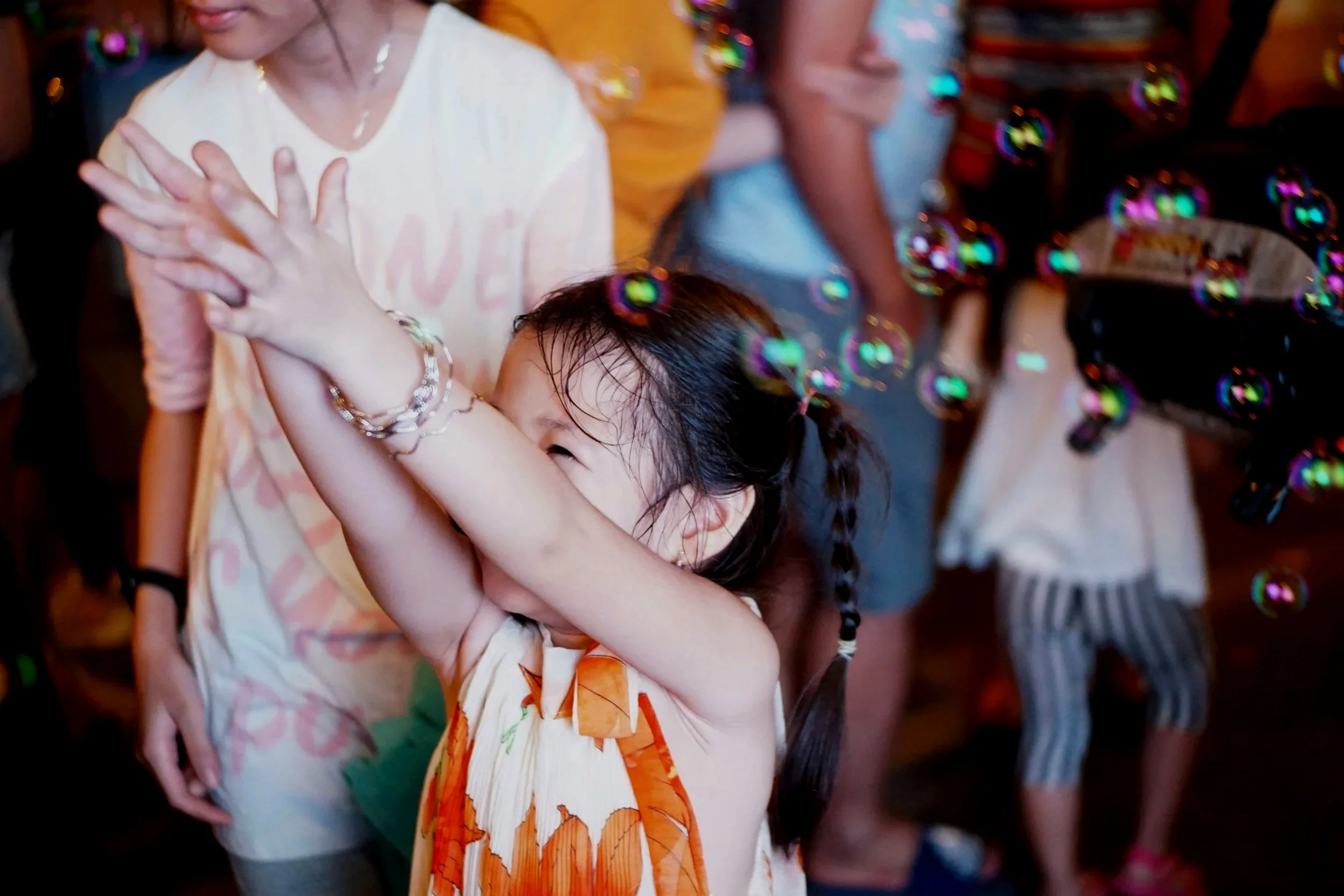 A young girl with braided hair wearing an orange and white dress is playing with soap bubbles at a party. An adult woman, in a light-colored dress with pink patterns, stands beside her. Other children and adults are visible in the background.
