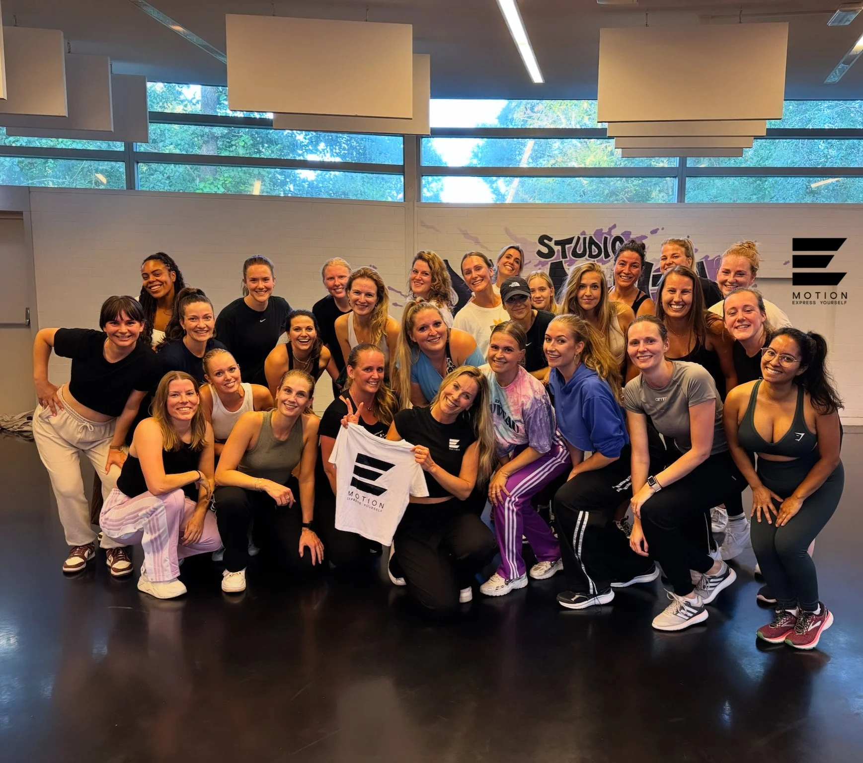 Group of women in athletic clothing posing together in a fitness studio, smiling and holding a white towel with a logo. The studio has large windows, a white brick wall, and the logo 'STUDIO' in the background.