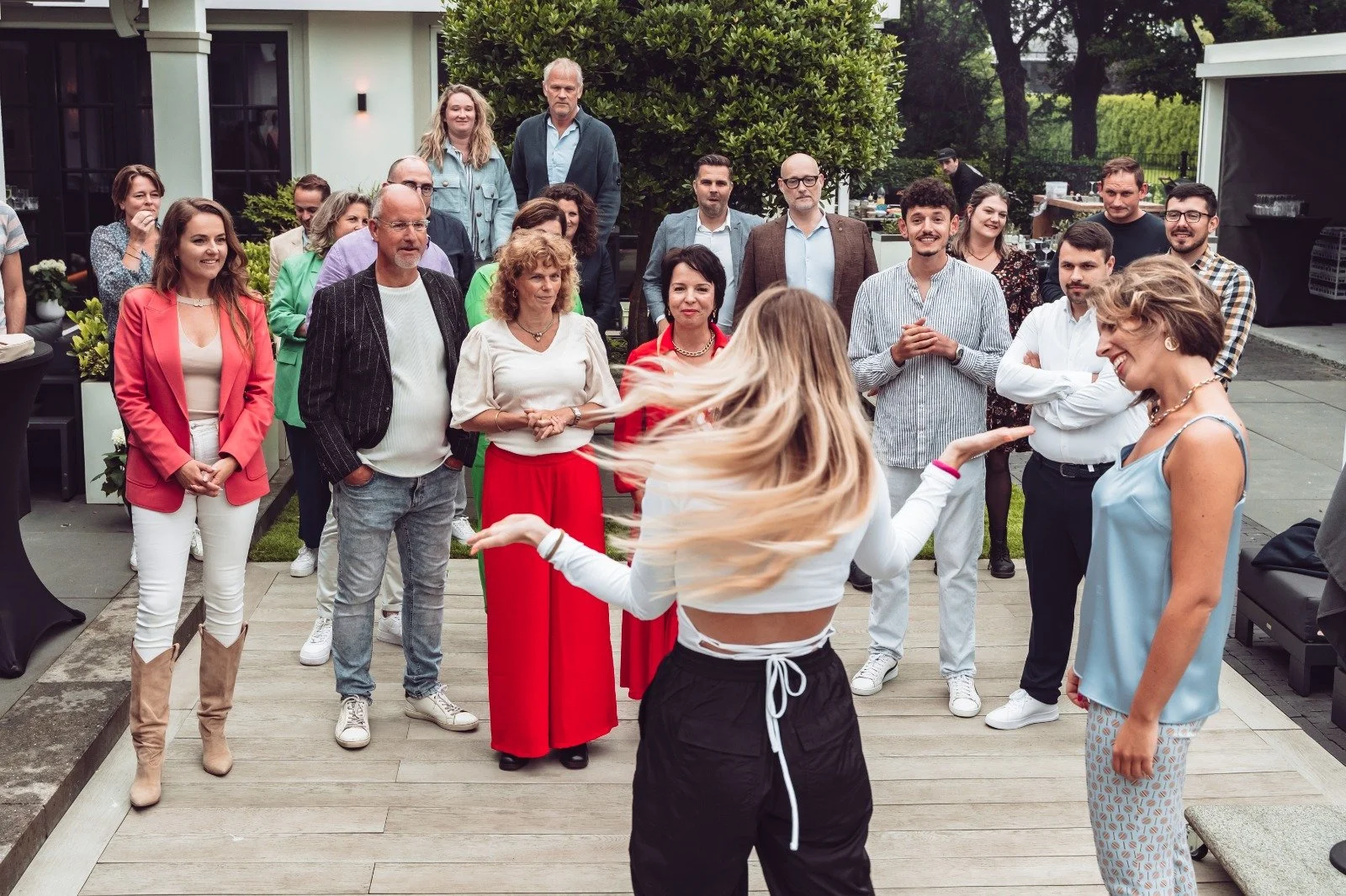 A woman with long hair dancing at an outdoor social gathering or party, with a group of people watching her and smiling.