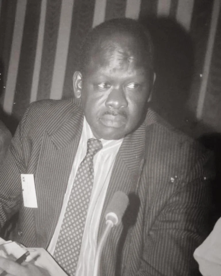 A black and white photo of a man in a suit and tie, sitting at a table with a microphone in front of him.