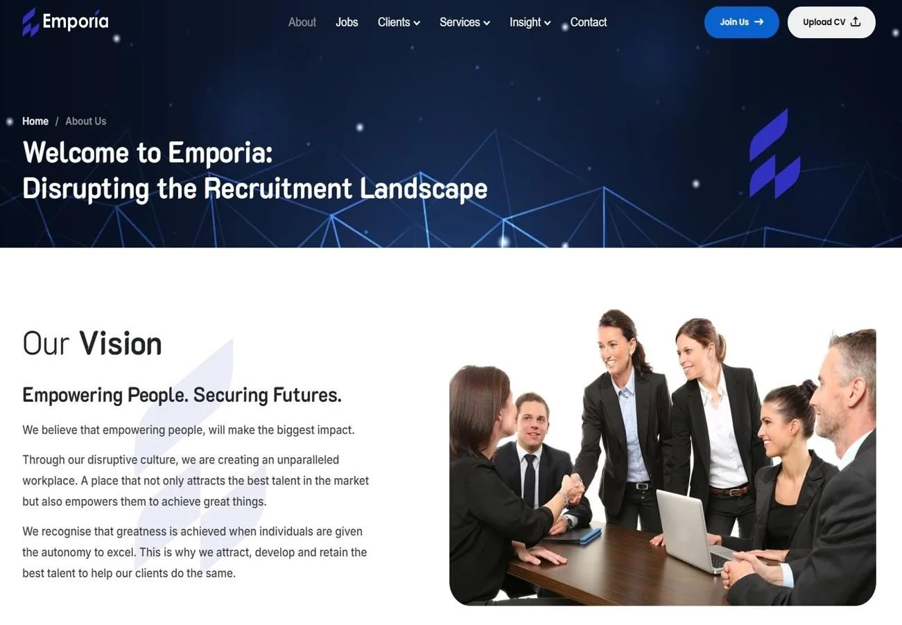 A group of five diverse business professionals, including three women and two men, engaged in a meeting around a table. They are smiling, with one woman shaking hands with another person across the table. The setting is a formal office with a laptop and notebooks on the table, indicating a collaborative business discussion.