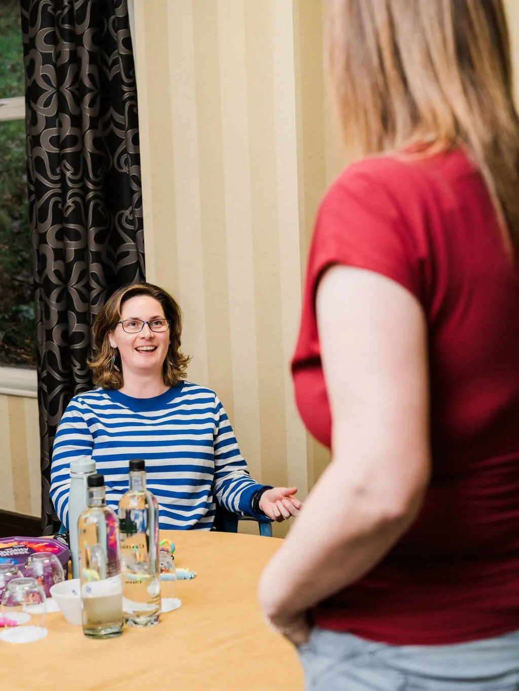 A woman in a blue and white striped shirt talking to another woman in a red shirt, seated at a table with water bottles, glasses, and snacks, indoors near a window with patterned curtains.