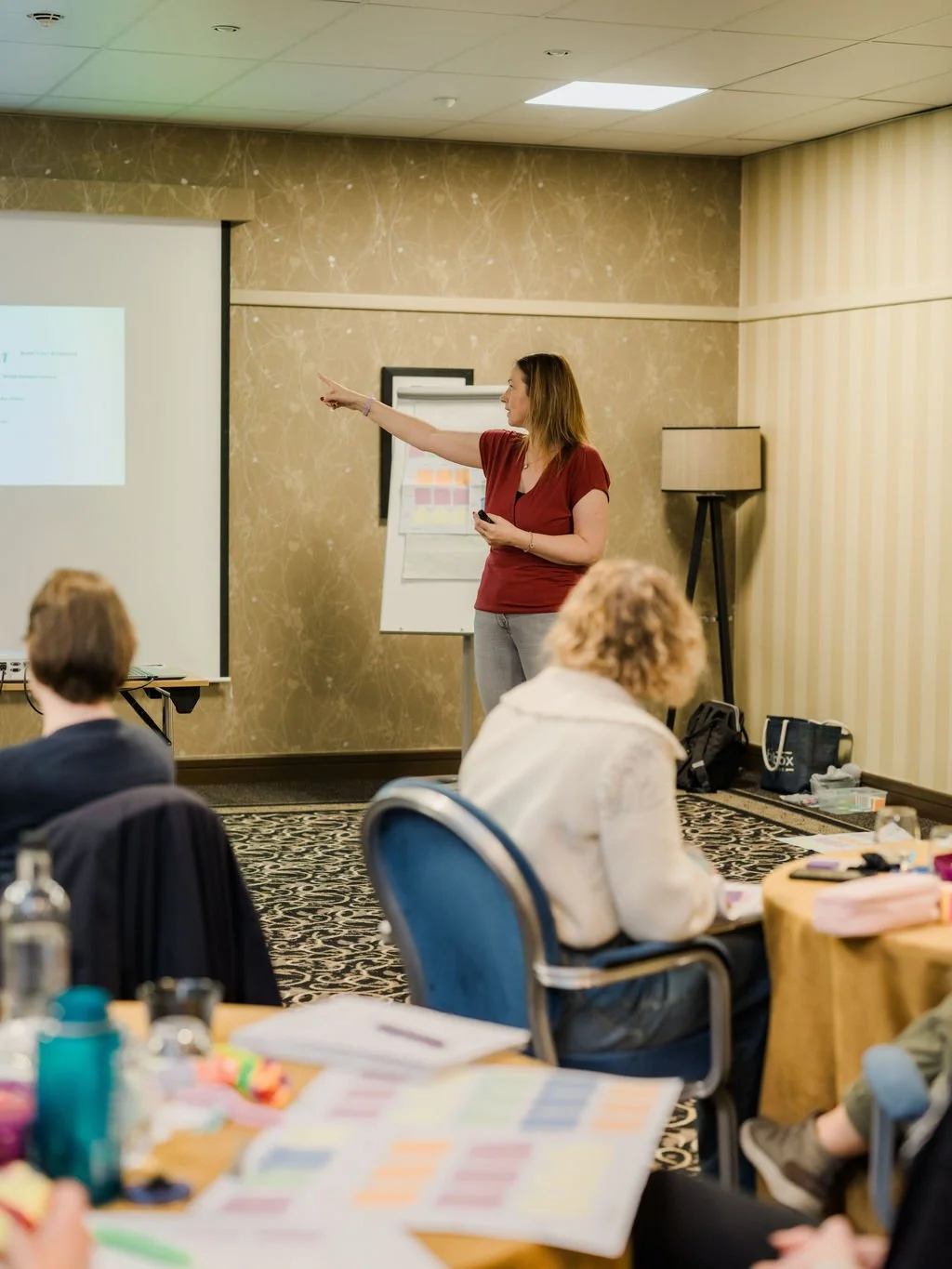 A woman in a red shirt giving a presentation to a group of people in a conference room, pointing at a screen.