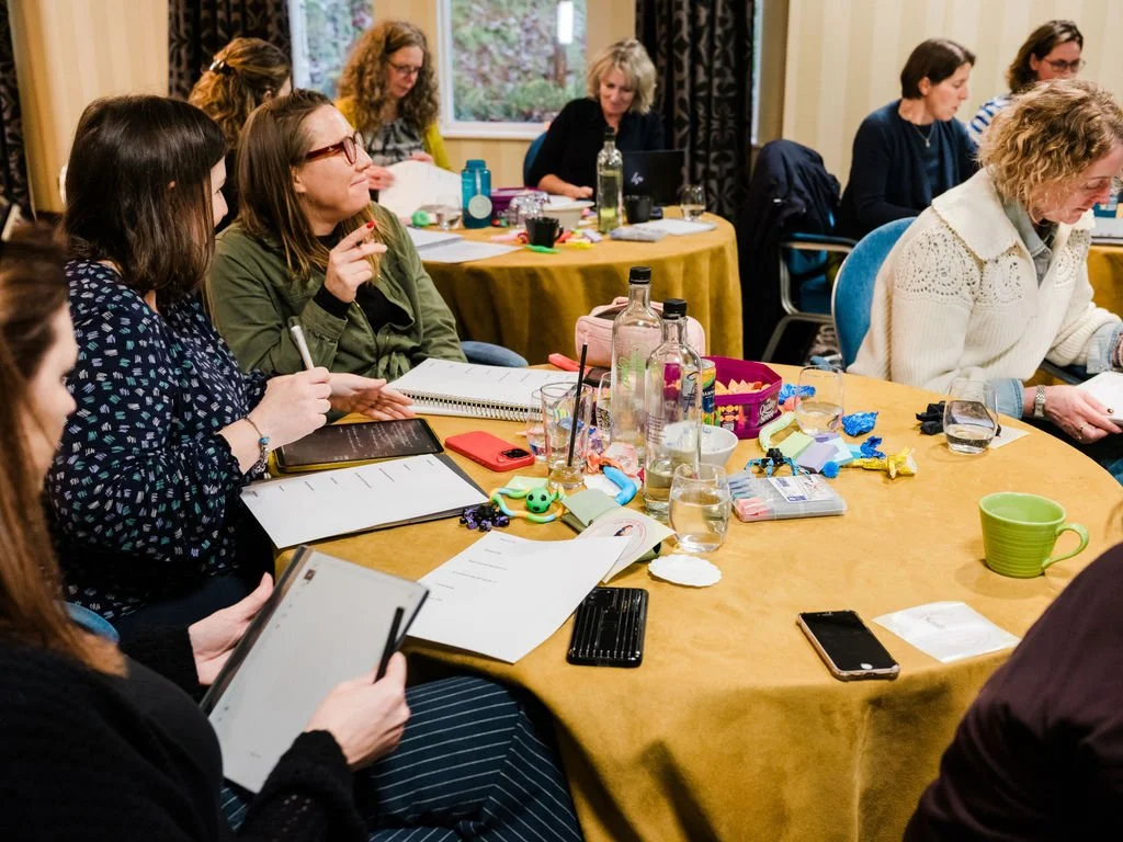 Women gathered around a table in a meeting or workshop, some taking notes and others listening. The table has water bottles, glasses, and various personal items.