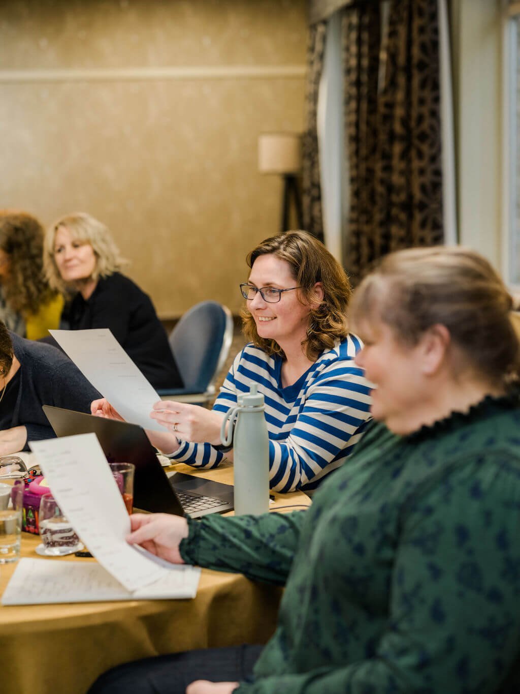 A group of women sitting at a conference table during a meeting, holding papers, with laptops and drinks on the table.