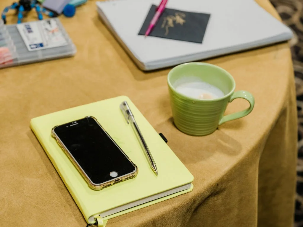 A yellow notebook with a smartphone and a silver pen on top, a green mug with a beverage, a white laptop, a pink pen, and a notepad on a table.