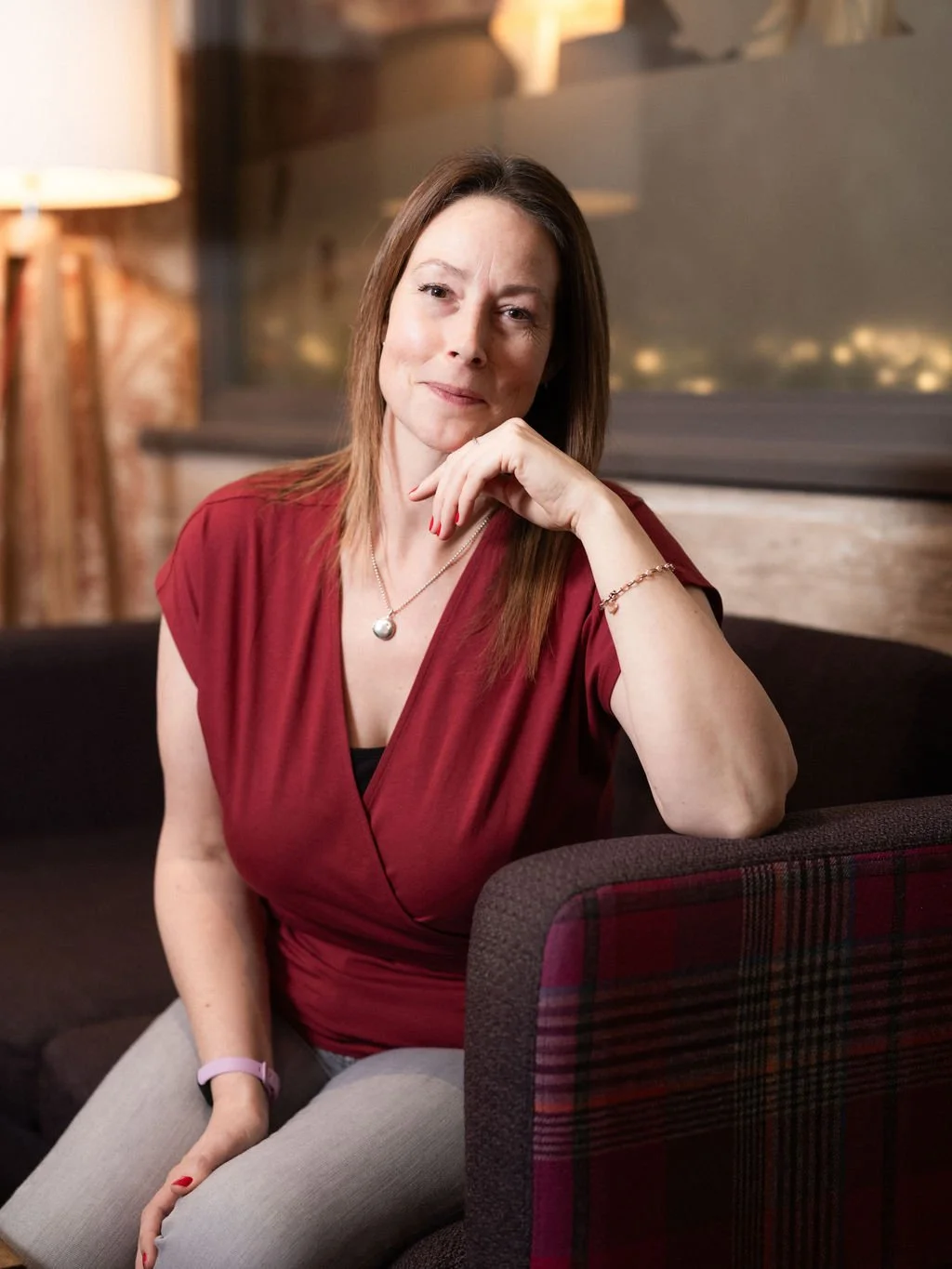 A woman with shoulder-length brown hair, wearing a red top and gray pants, sitting on a dark-colored sofa in a warmly lit room with wood and brick decor, resting her chin on her hand, looking at the camera.