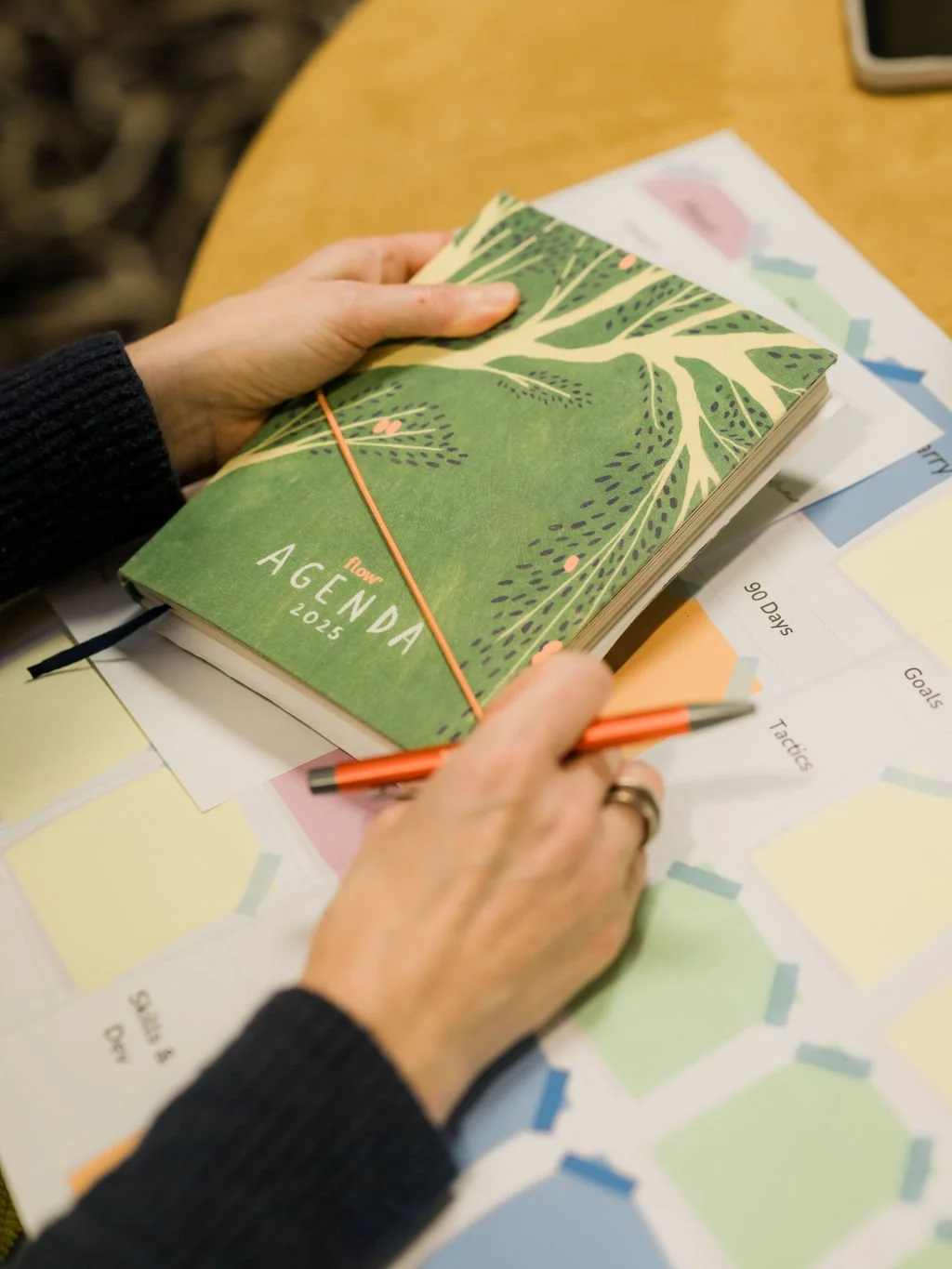 Hands holding a green, illustrated 2025 agenda planner with orange elastic band, on a wooden table with papers and a pen.