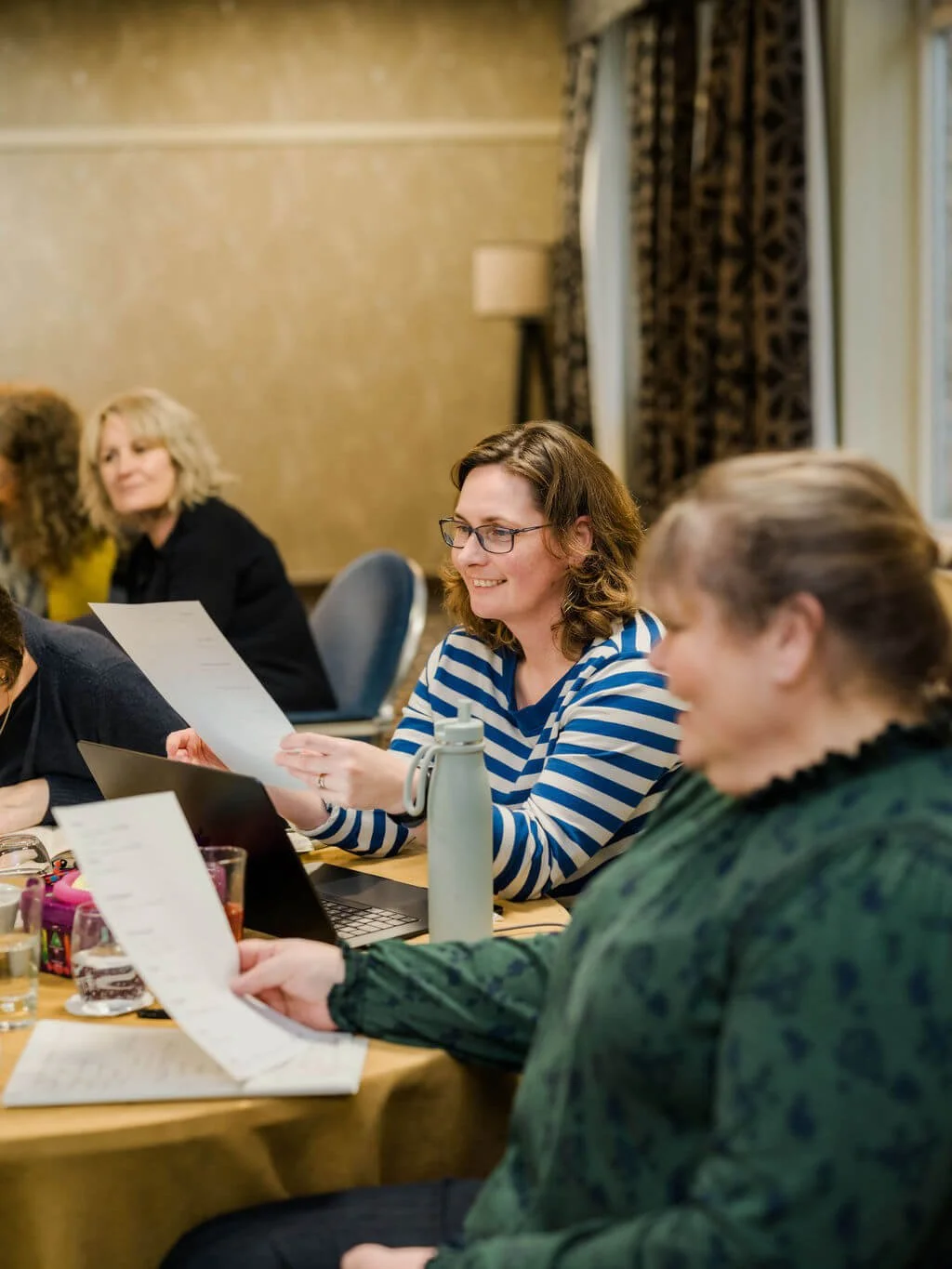 Group of women sitting at a conference table, reading papers, in a room with patterned curtains and warm lighting.