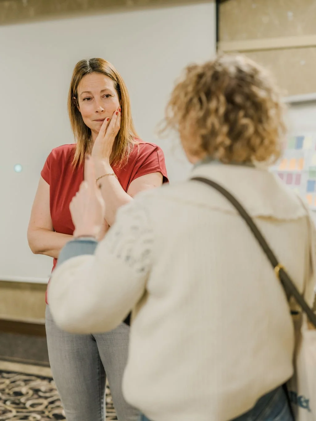 Two women in a conversation, one woman with a confused expression touching her face, and the other woman with curly hair, gesturing with her hand.
