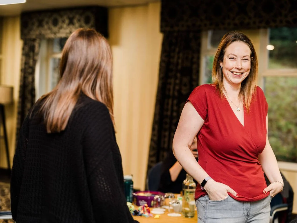 Two women talking and laughing indoors, with a table of supplies or crafts in the background, and windows with curtains.