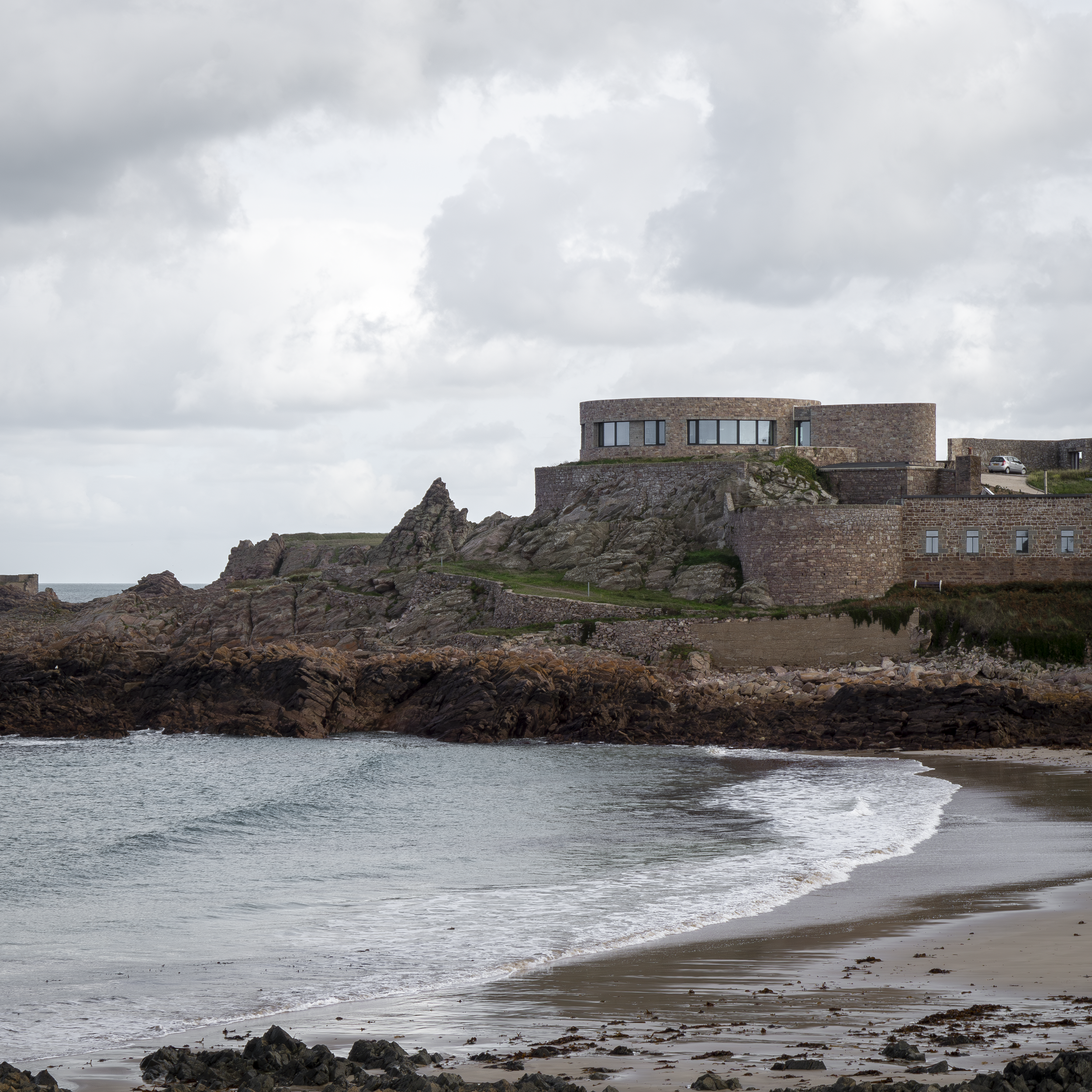 Coastal scene with a modern stone building on a rocky hill overlooking the ocean, cloudy sky, and sandy beach in the foreground.
