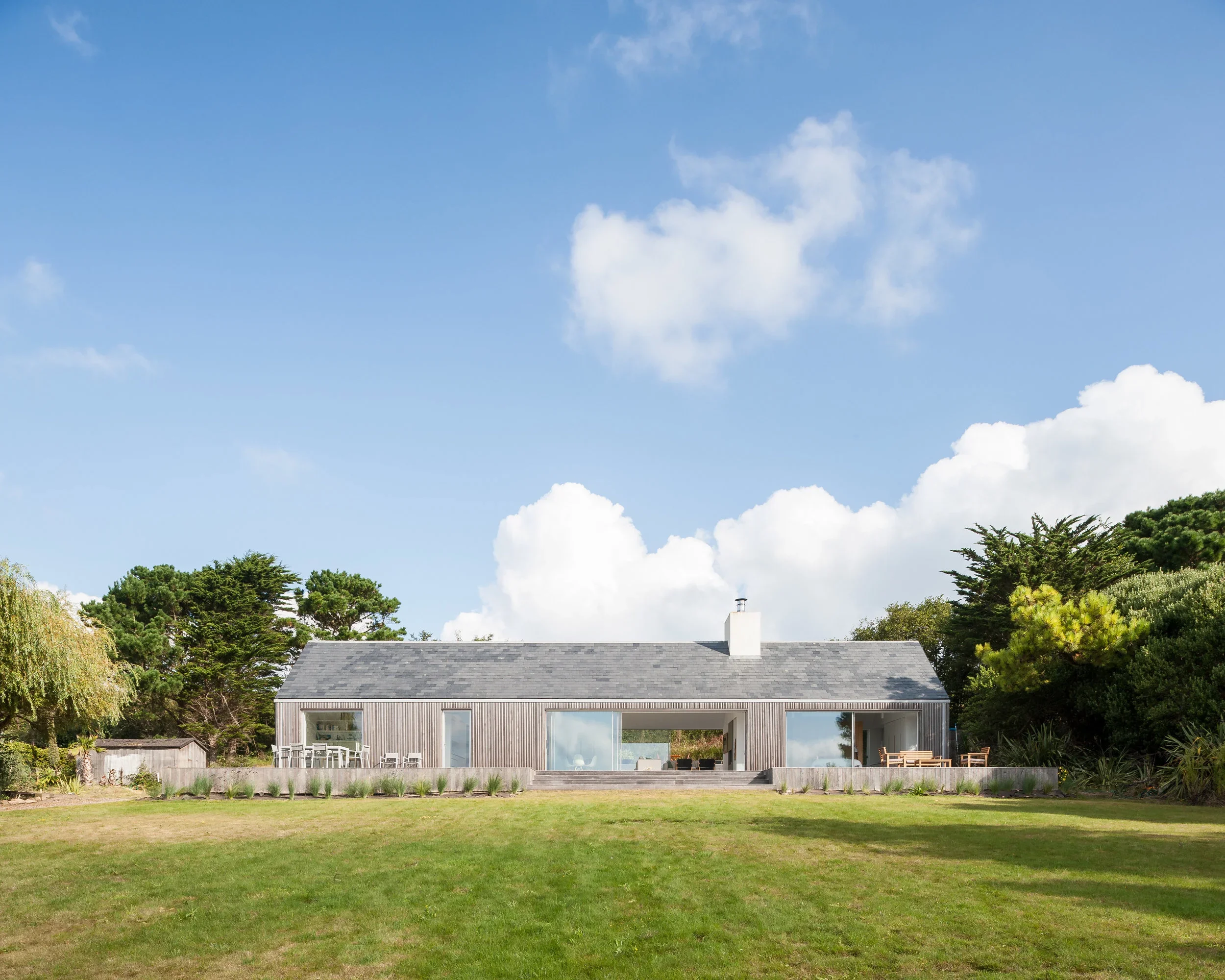 A modern house with a sloped roof, large glass sliding doors, and patio furniture, set in a lush green yard with trees and a blue sky with white clouds.