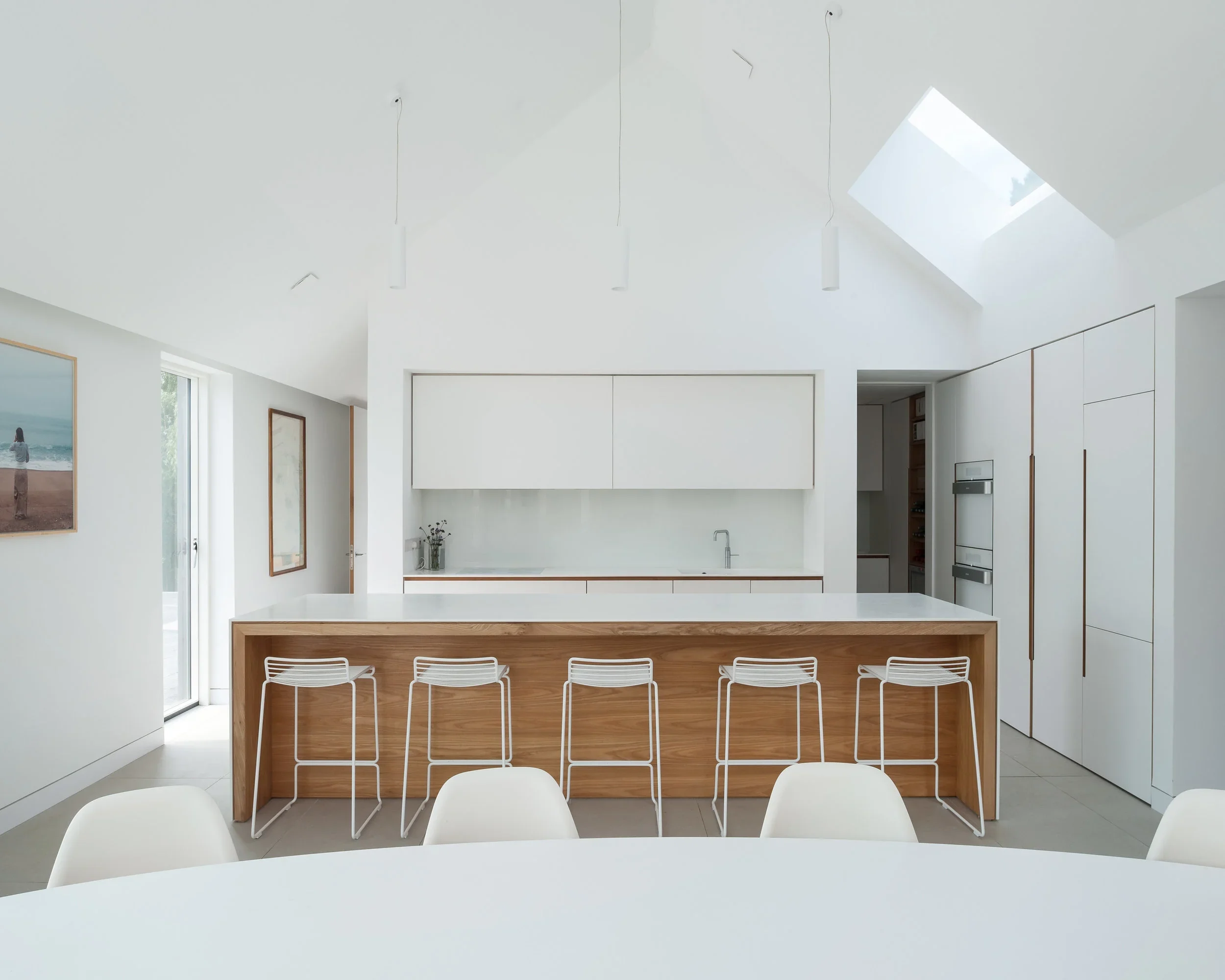 Modern white kitchen with wooden accents, high ceiling, skylight, island with white countertop, six white bar stools, and artwork on the walls.