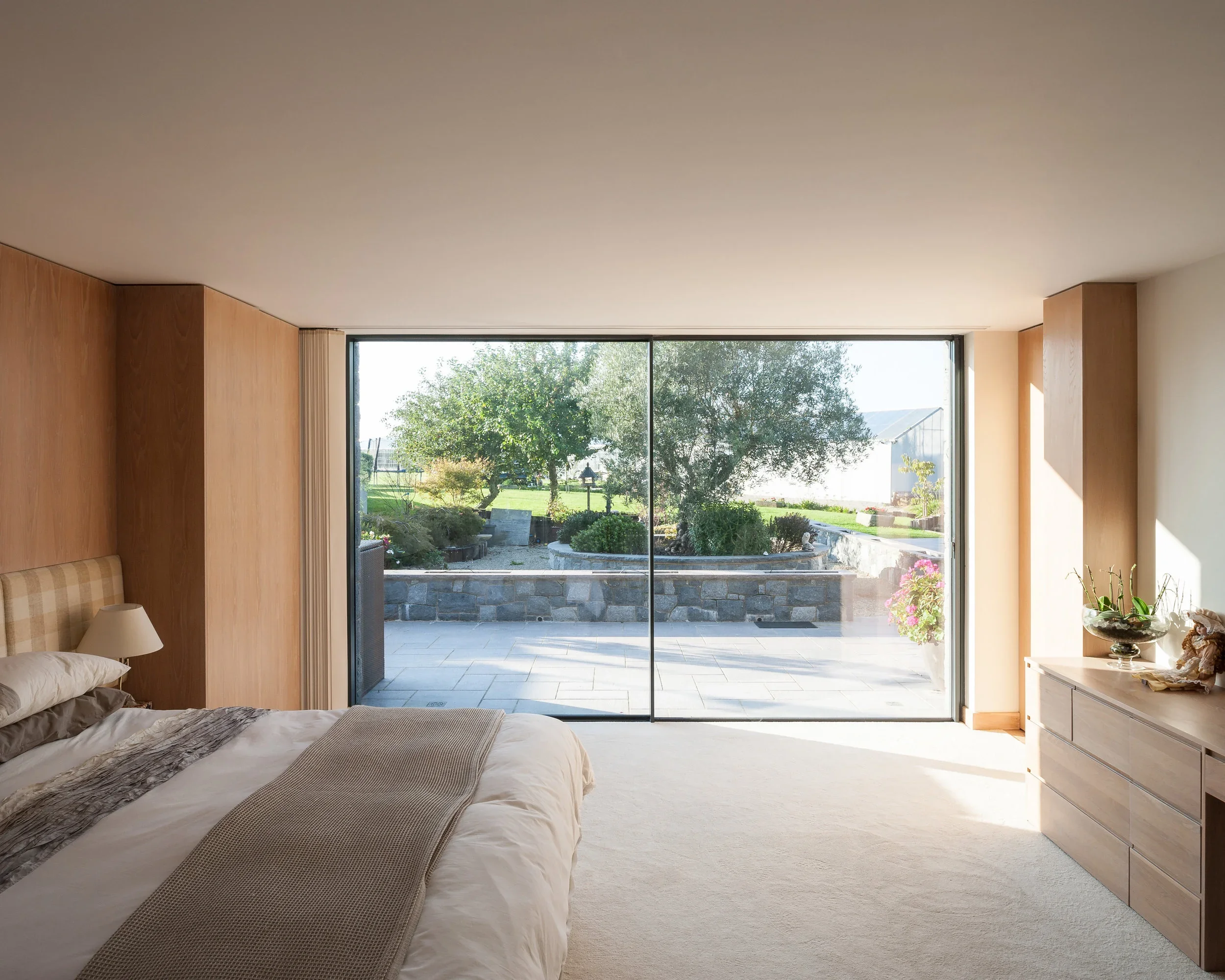 Bedroom with large glass sliding doors opening to a patio and garden outside. The room has a bed with a beige headboard, beige and white bedding, a nightstand with a lamp, and a dresser with decorative items.