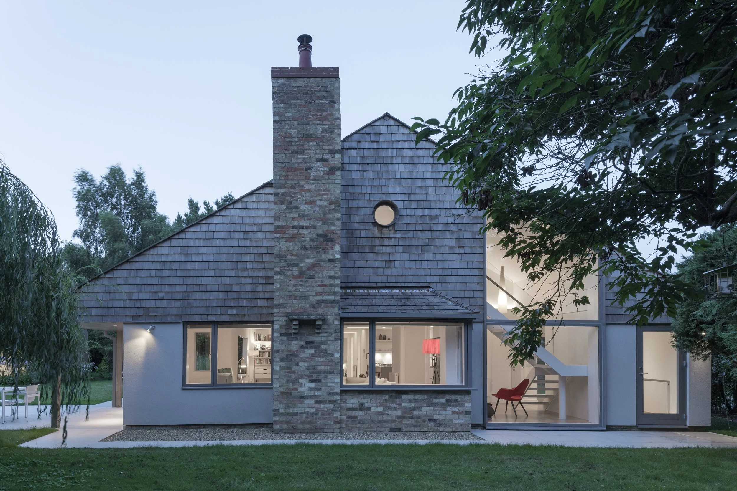Modern two-story house with large windows, brick chimney, and sloped roof. Interior lights visible through the windows, with a staircase and a red chair inside. Trees and a grassy yard surround the house.