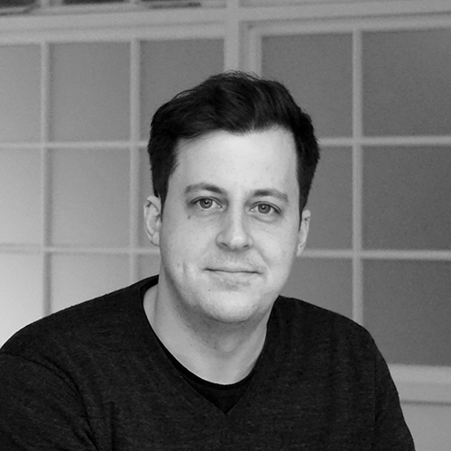 Max Babbe - Black and white portrait of a man with short dark hair wearing a dark shirt, sitting in front of a windowed background.
