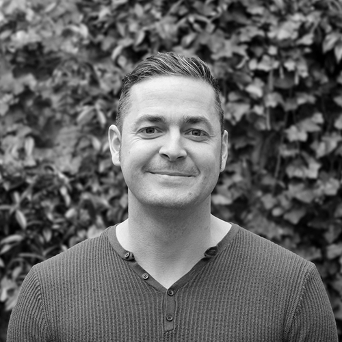 Jamie Le Gallez - A man with short hair, wearing a ribbed henley shirt, smiling in front of leafy bushes.
