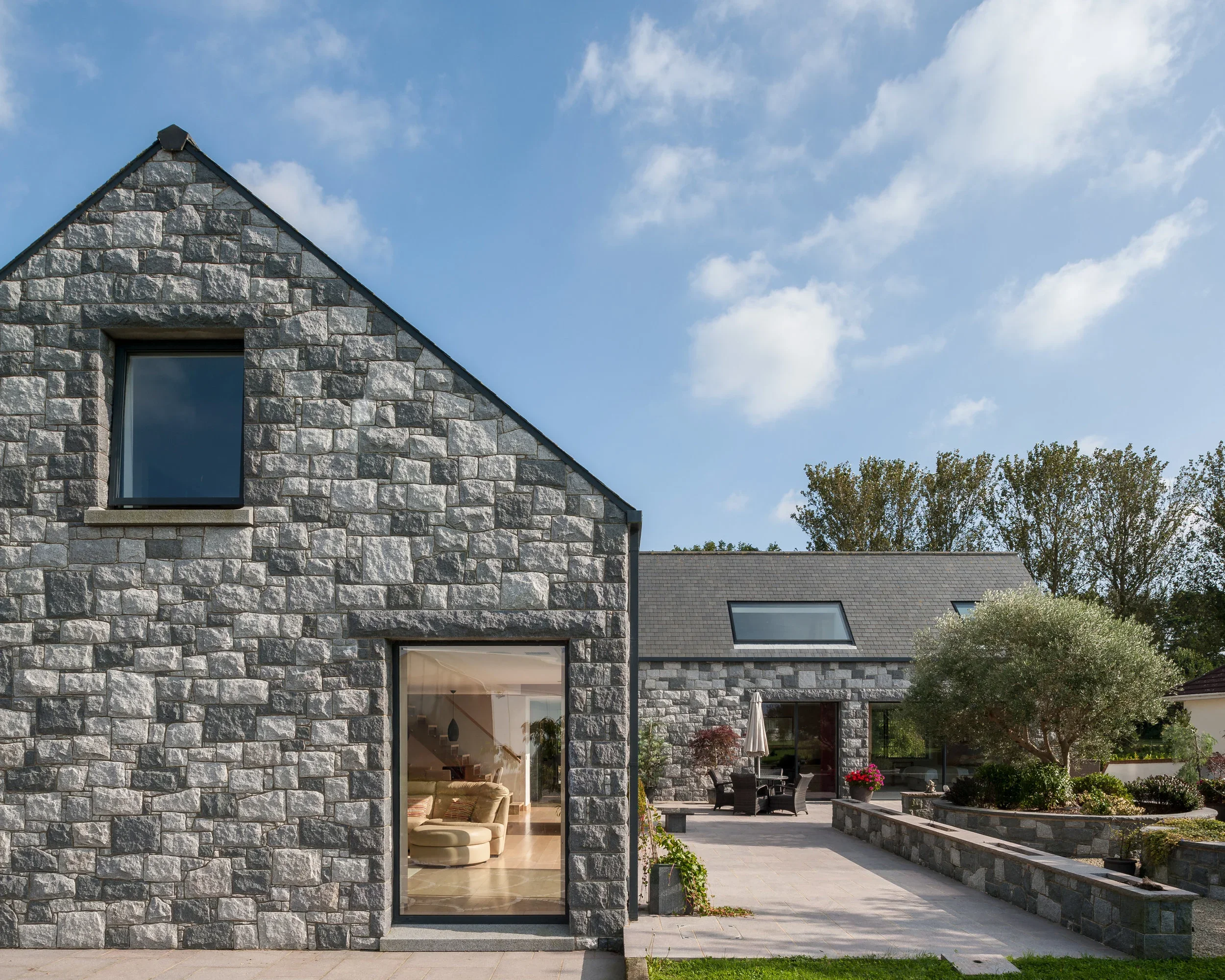 Modern stone house with large windows, outdoor patio area, and landscaped garden under a blue sky with scattered clouds.