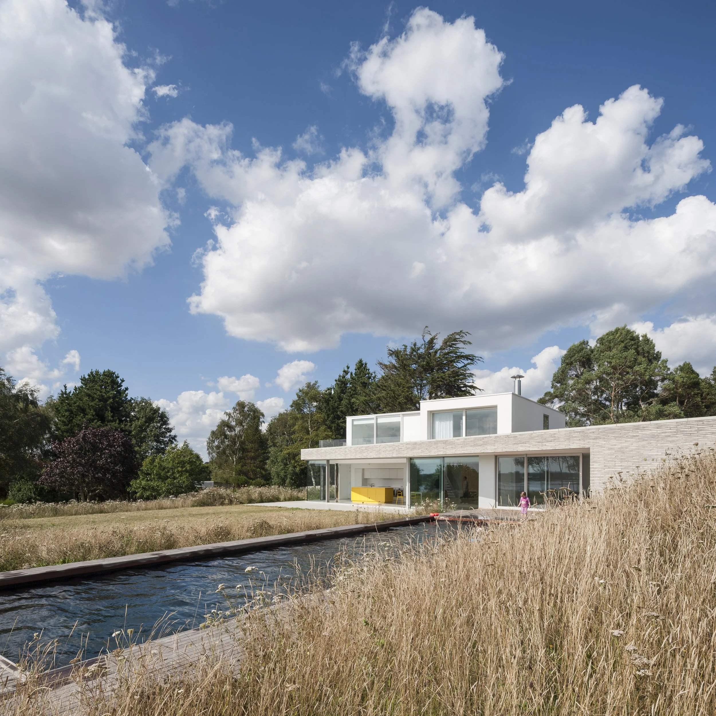 Modern white house with large glass windows surrounded by tall dry grass, trees, and a blue sky with scattered clouds.