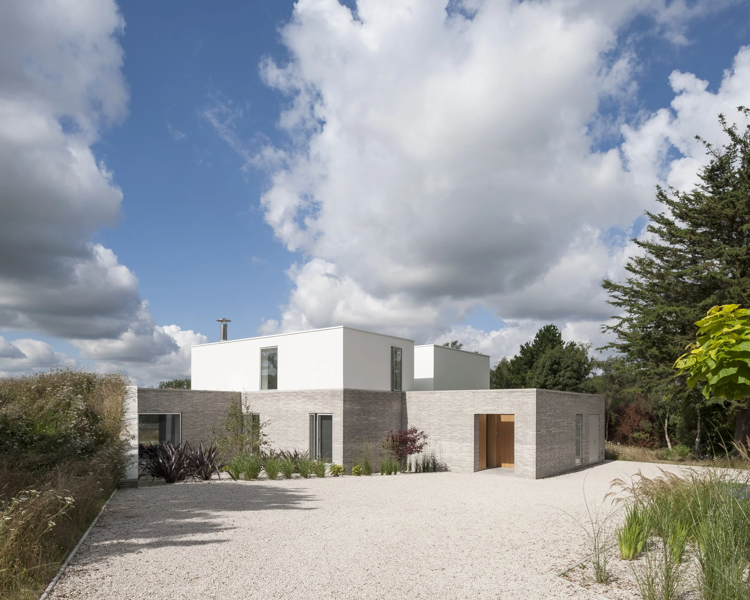 Modern house with white and brick exterior, surrounded by landscaped garden and gravel driveway under a partly cloudy sky.