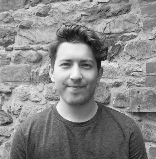 Michal Hart - A young man with wavy hair and a light beard smiling in front of a stone wall, wearing a dark T-shirt.