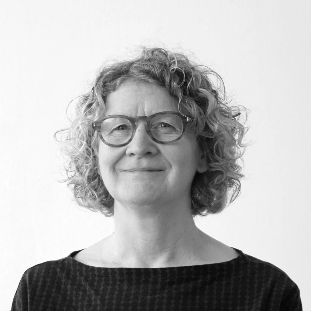 A man with glasses and a beard in front of leafy bushes.