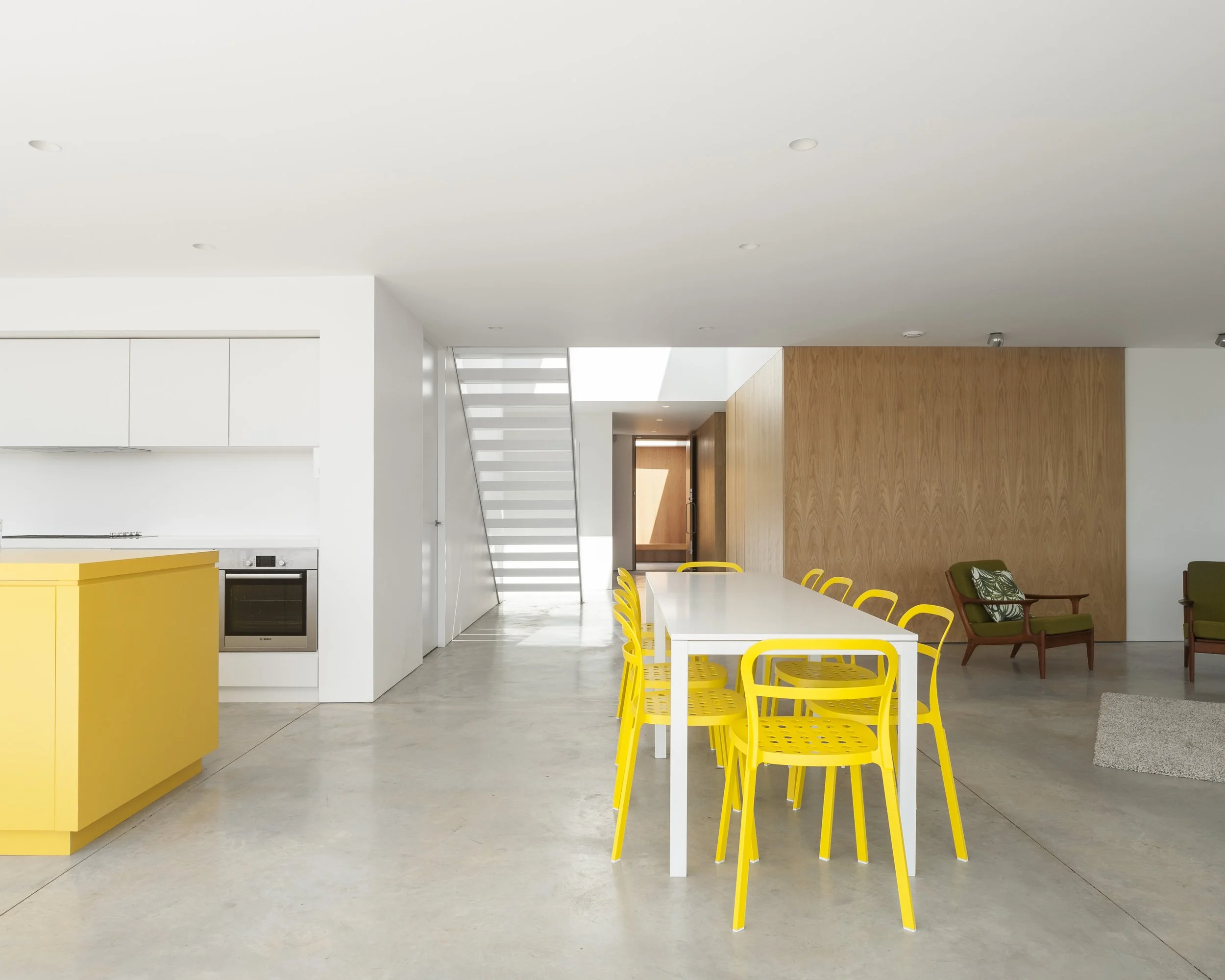 Modern open-concept kitchen and dining area with yellow chairs around a white table, a yellow kitchen island, and green armchairs with a wooden accent wall.